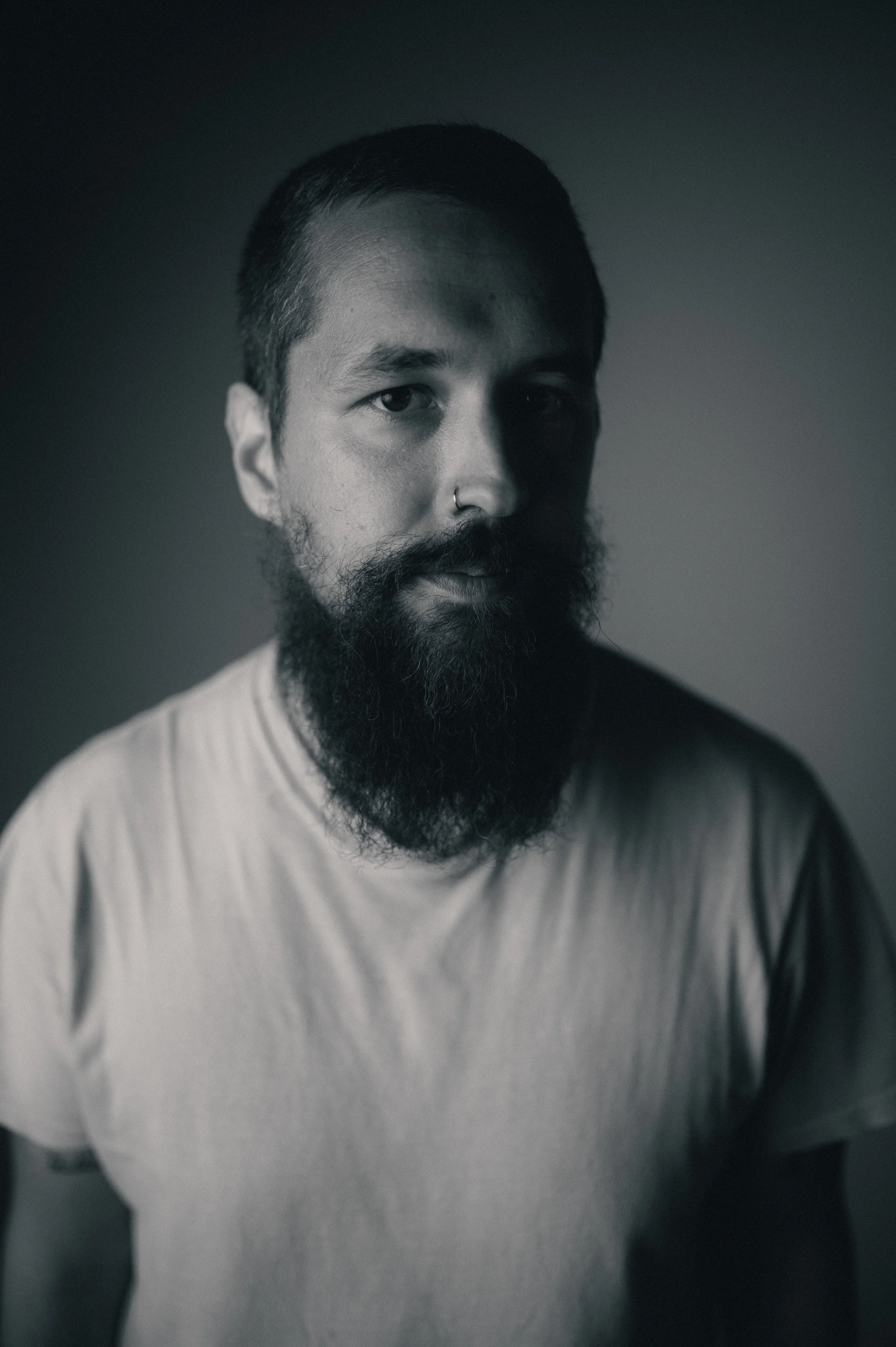 Black and white portrait of a bearded man with a nose ring, wearing a light-colored T-shirt, looking directly at the camera against a dark gradient background.