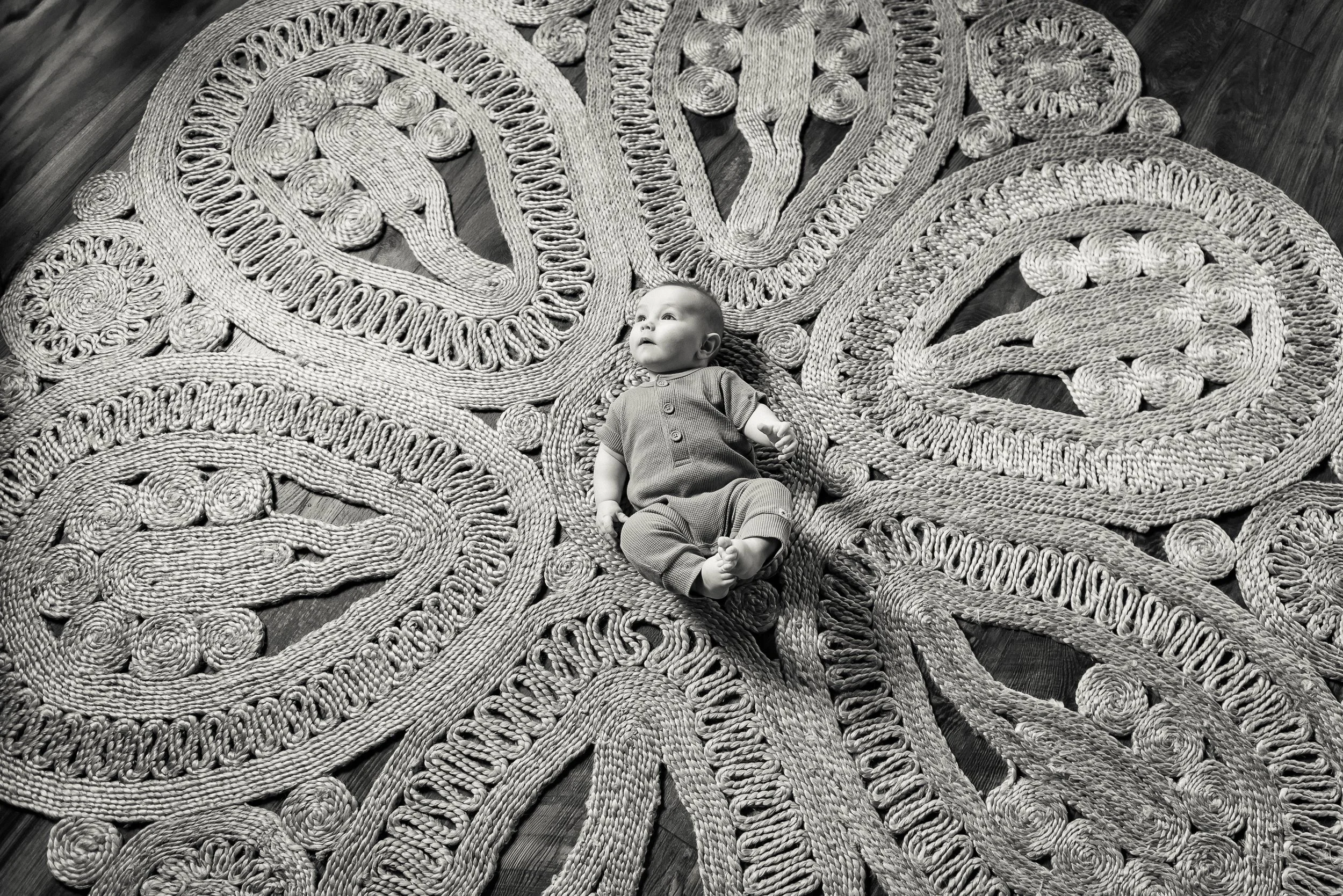 A baby lying on a large, intricately woven, decorative rug on a hardwood floor.