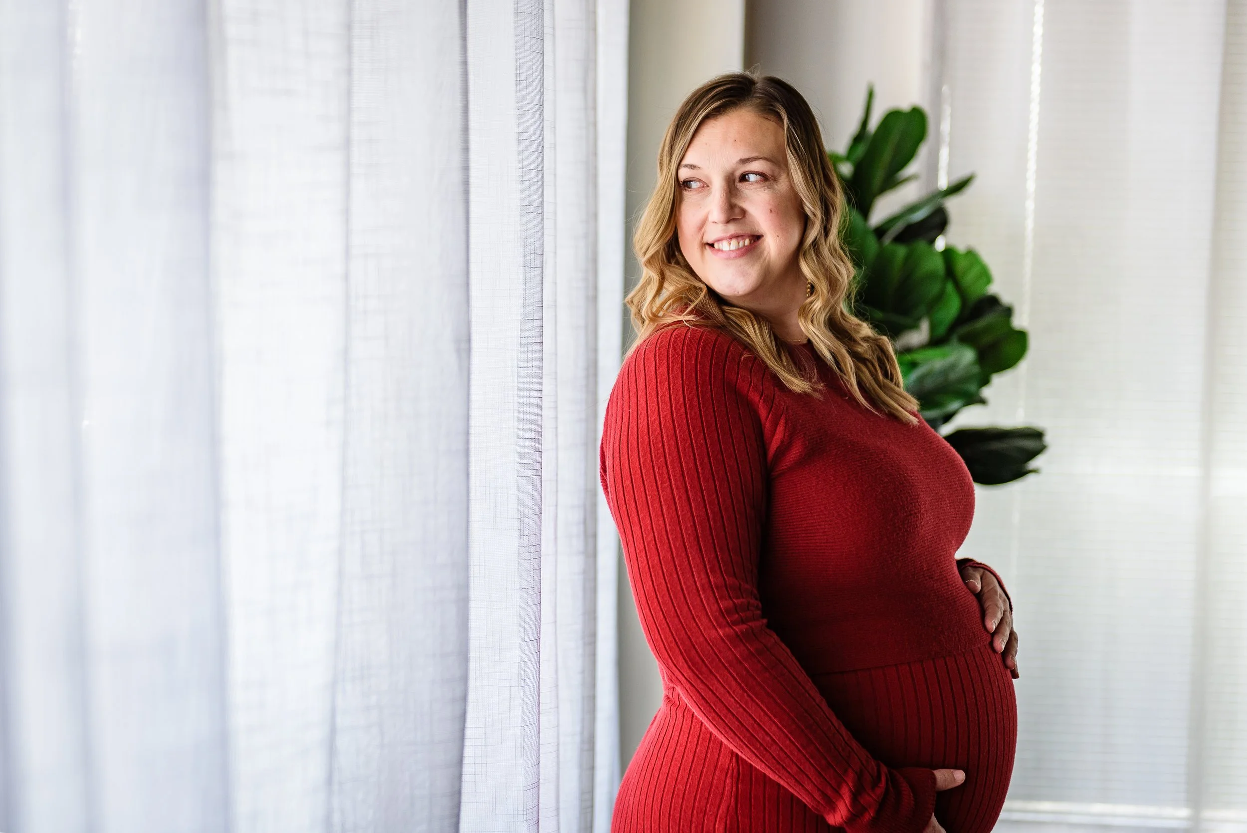 A pregnant woman with blonde hair wearing a red long-sleeve sweater standing indoors near sheer curtains, smiling and looking to the side, with a green plant in the background.