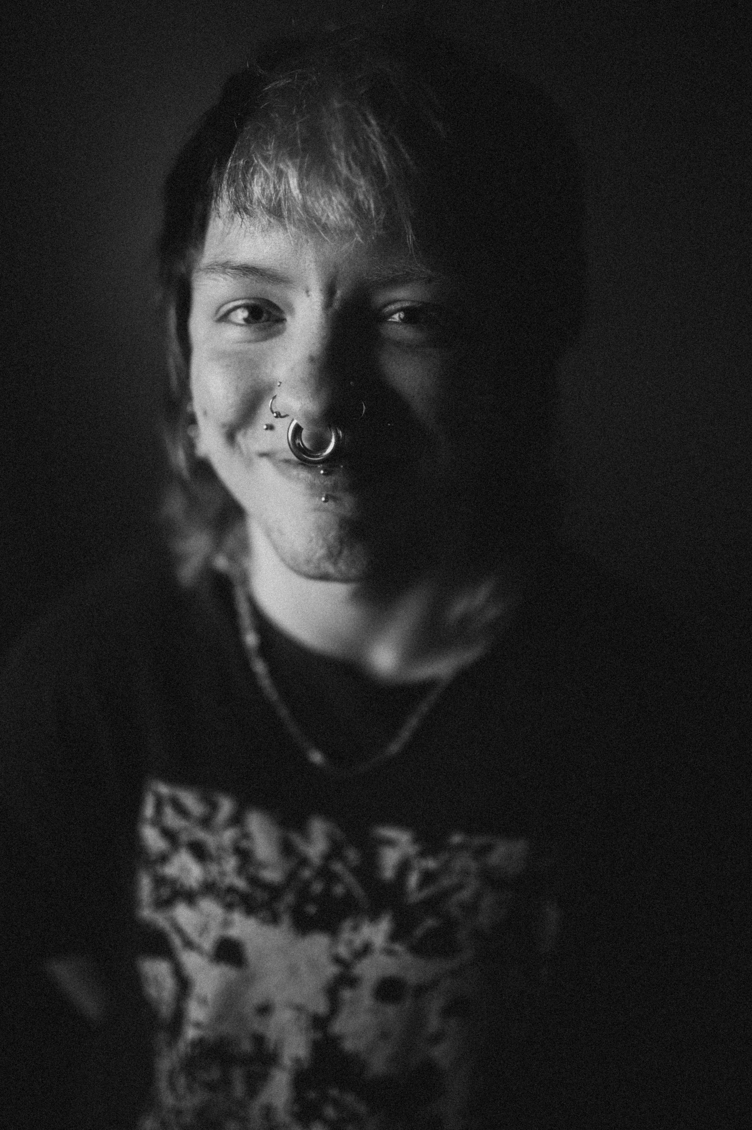 Black and white close-up portrait of a person with short hair, multiple facial piercings, and a slight smile, against a dark background.