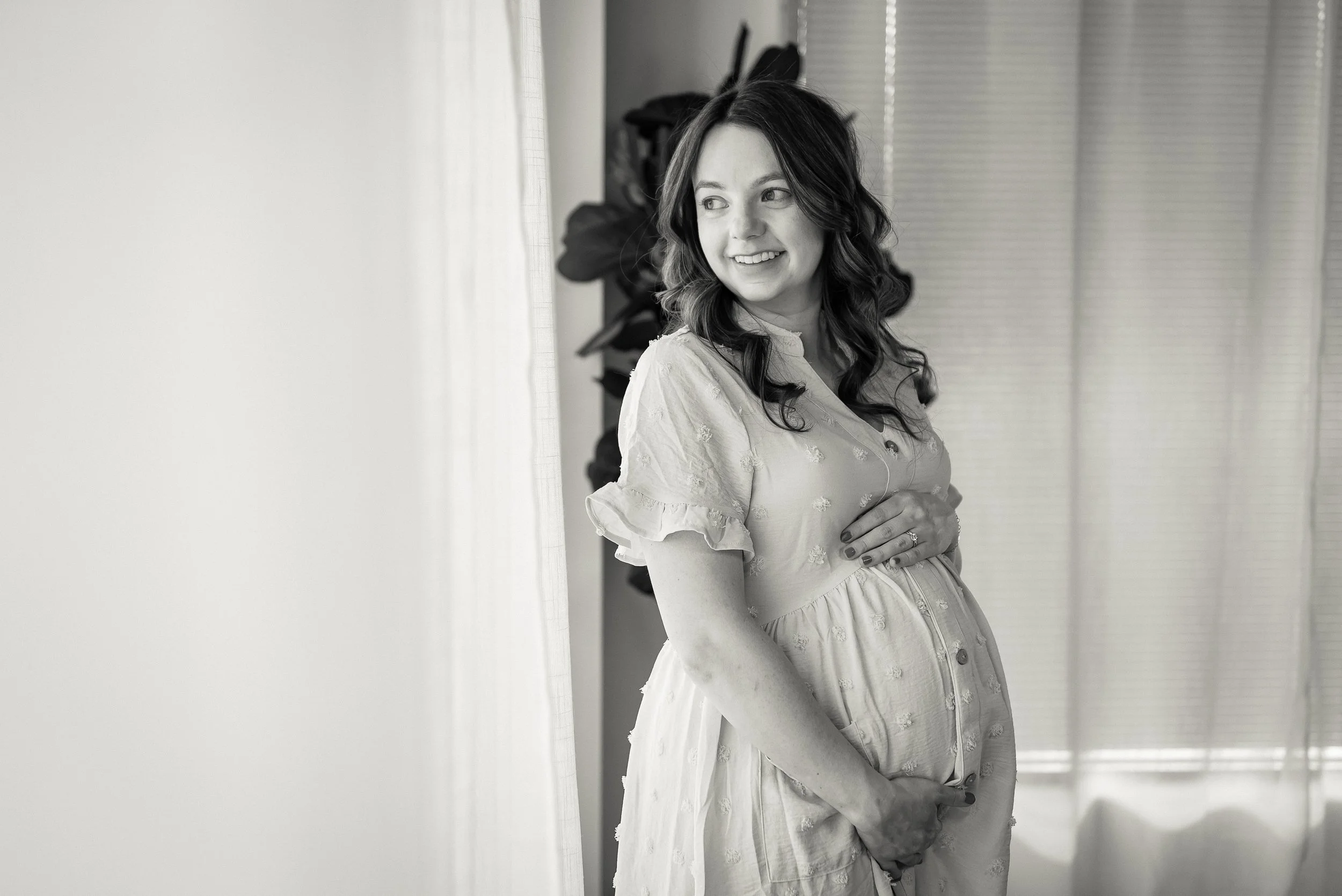 A pregnant woman standing indoors, smiling, with one hand over her belly and the other holding her belly from below, near a window with curtains.