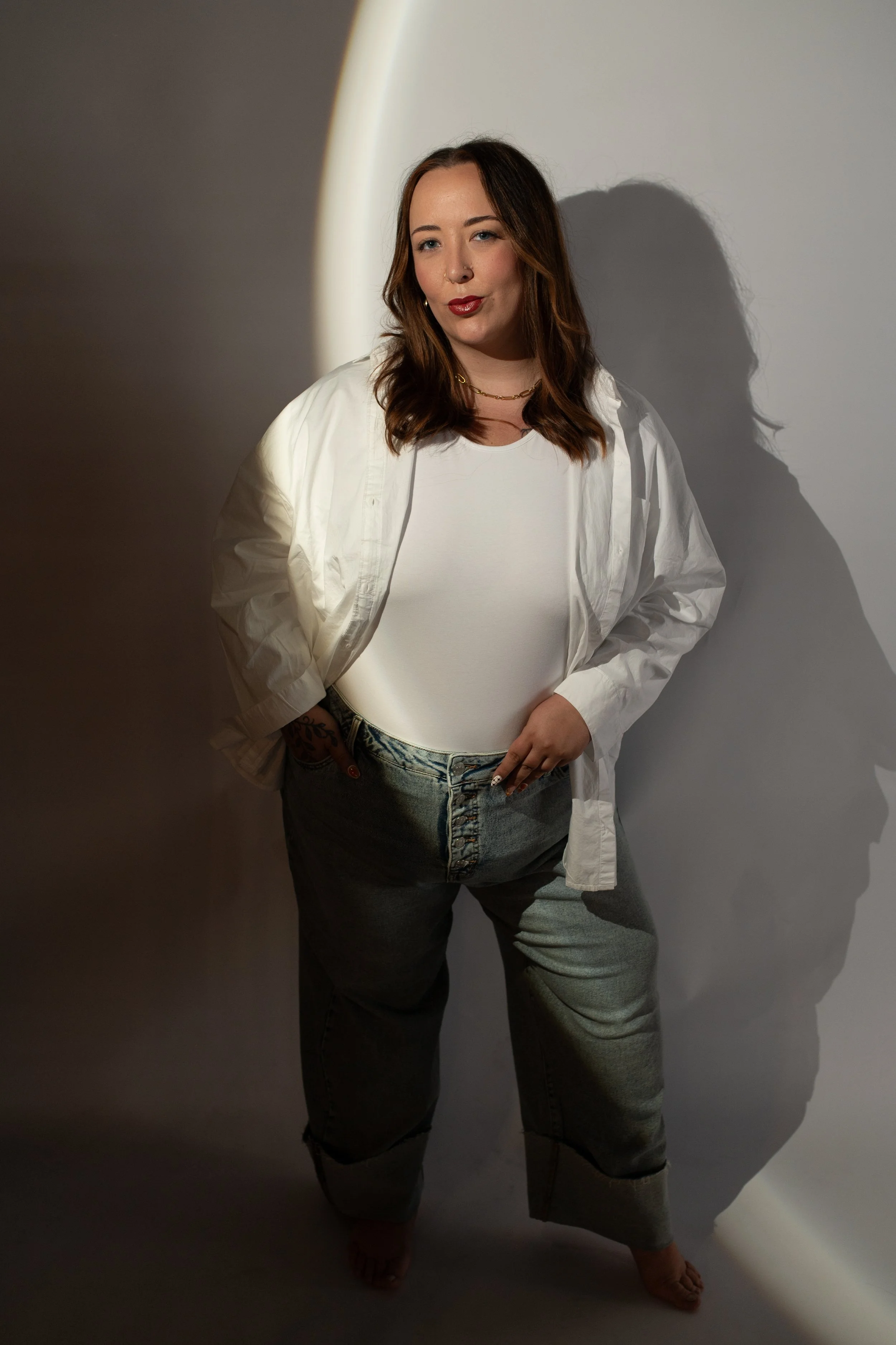 A woman with long brown hair, wearing a white t-shirt, white open shirt, and gray jeans, standing in front of a wall with a shadow cast behind her.