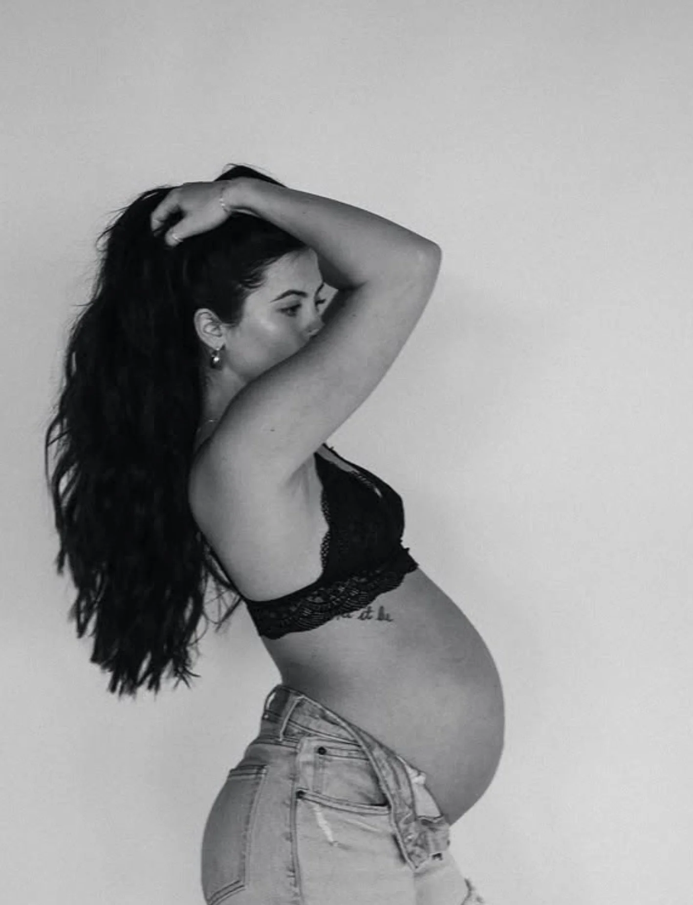 A pregnant woman with long, dark hair, wearing a black lace bralette and light-colored jeans, is posed against a plain wall, with one arm raised and her hand resting on her head, showing her side profile.