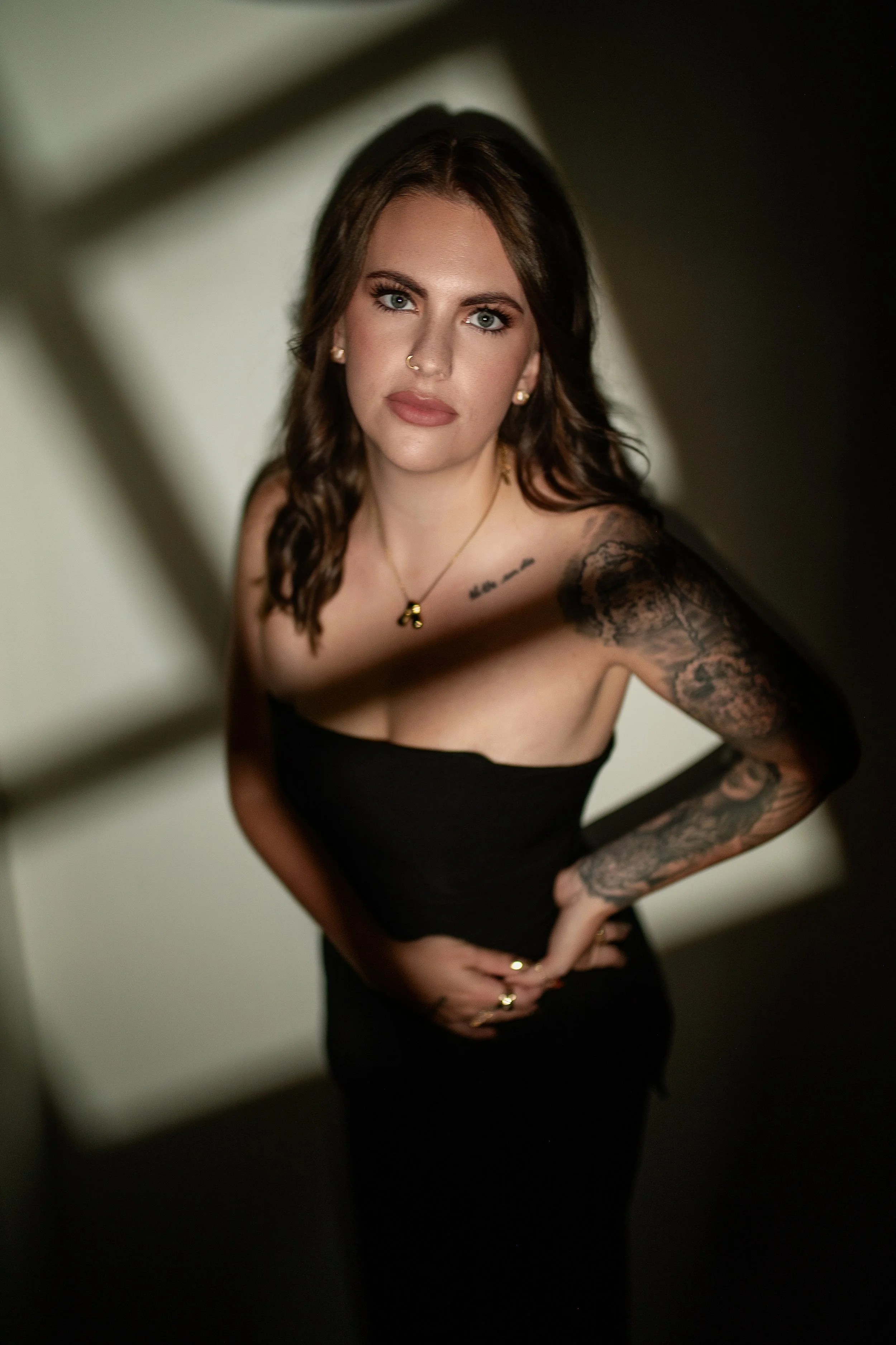 A woman with long, wavy brown hair and blue eyes wearing a black strapless dress, gold jewelry, and tattoos on her right arm and chest, standing in a dimly lit corridor with her hand on her hip.