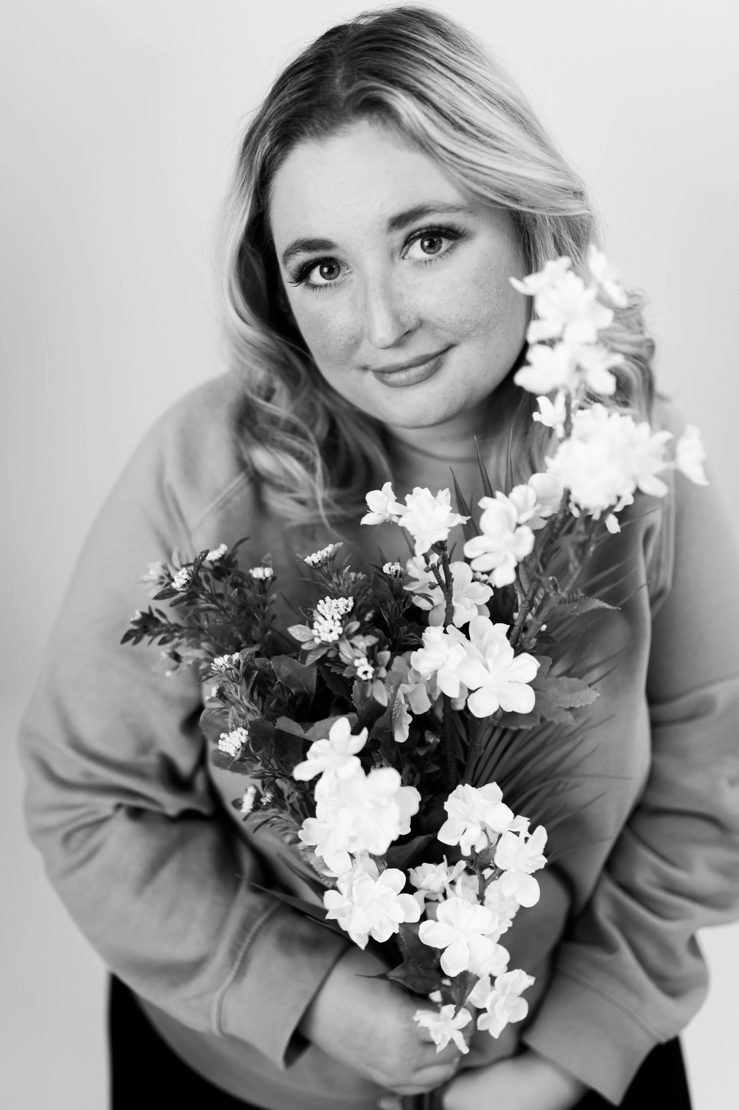 Black and white portrait of a young woman with wavy hair holding a bouquet of flowers, smiling softly, wearing a casual jacket.