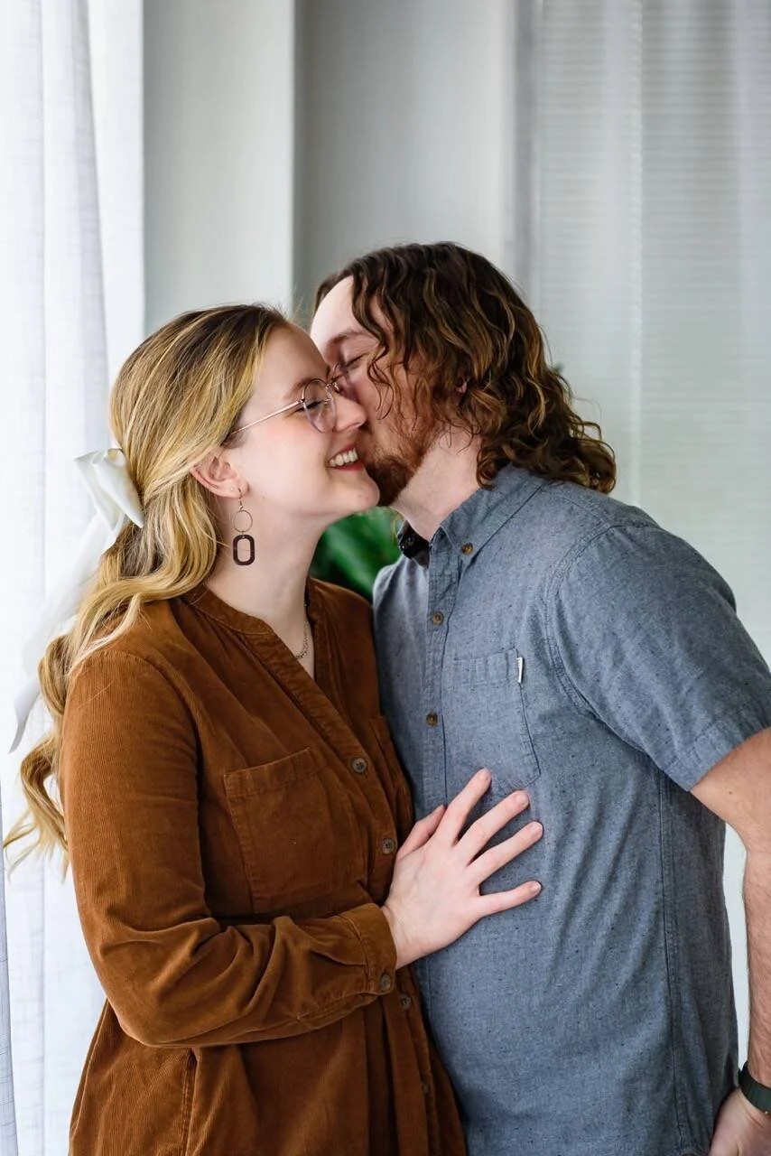 A couple with light skin and curly hair sharing a kiss, smiling, indoors near a window with white curtains.