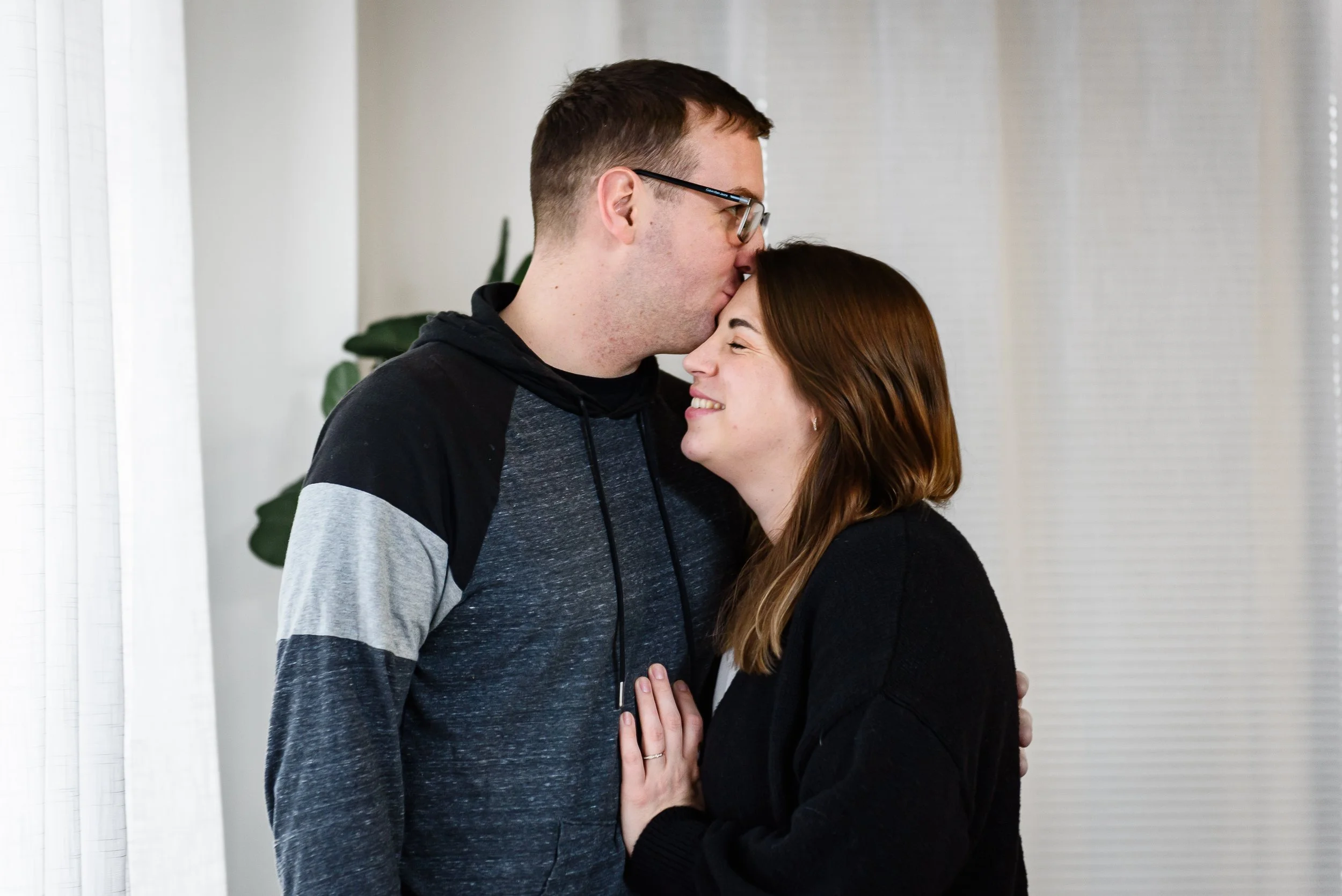 A man with glasses kissing a woman on the forehead as they smile and embrace indoors.
