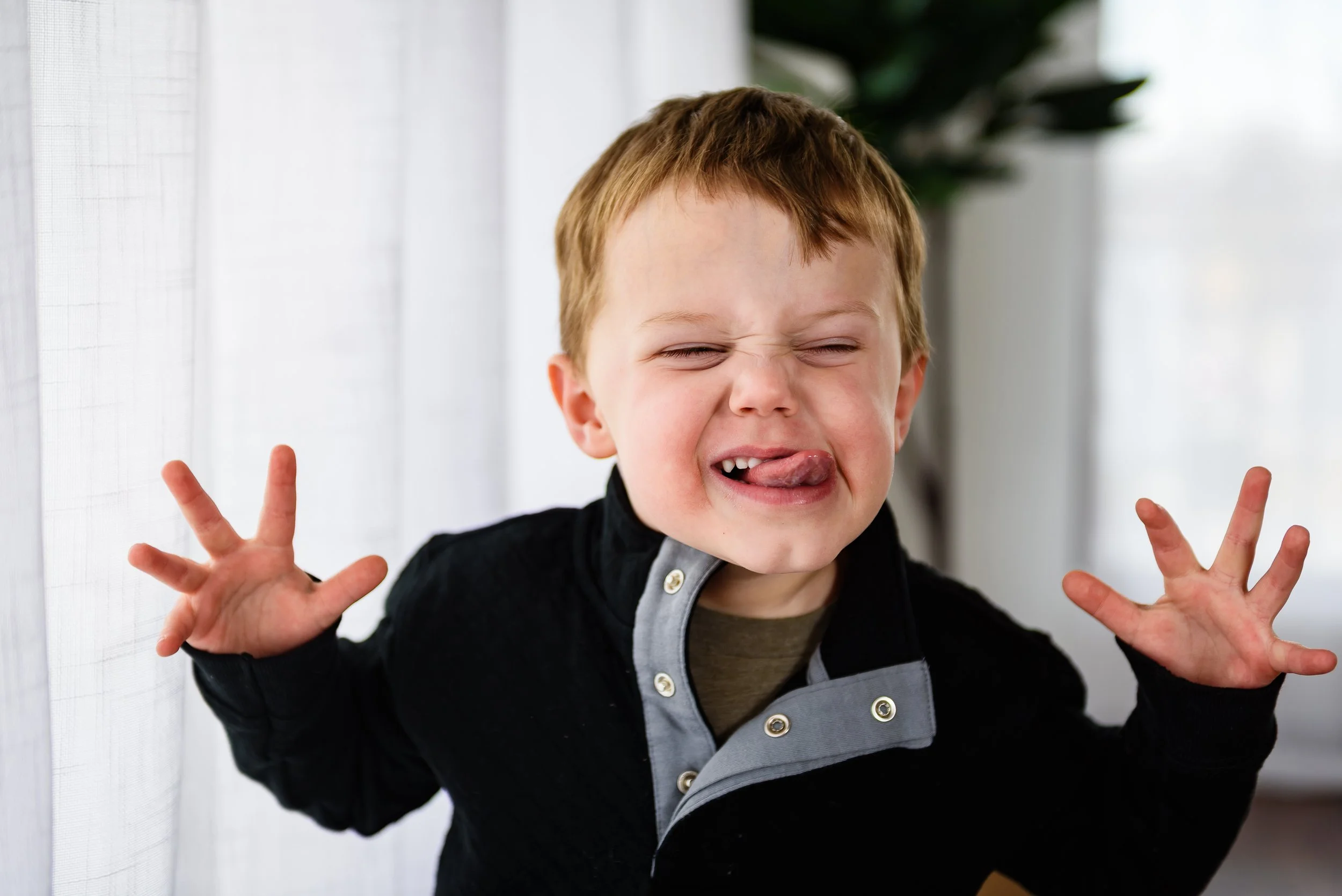 A young boy making a silly face with his eyes closed and tongue sticking out, hands raised with fingers spread, standing indoors near a window.