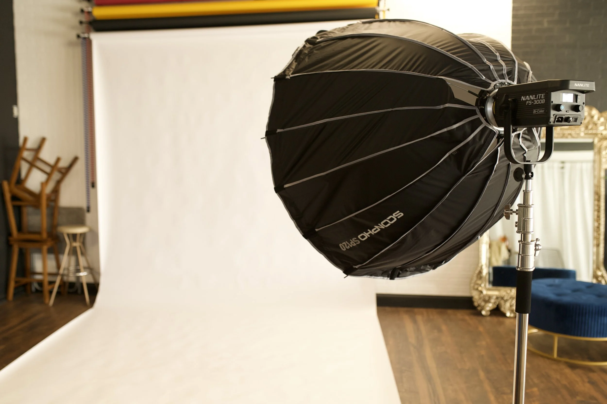 Studio photography setup with a large umbrella light modifier on a stand and a white backdrop in the background, with chairs and a mirror in the room.