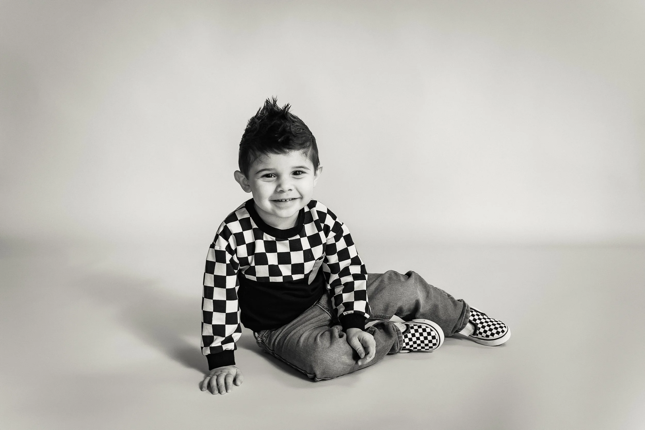 A smiling young boy with spiky hair, sitting on the floor against a plain background, wearing a checkered long sleeve shirt, gray pants, and checkered slip-on shoes in black and white.