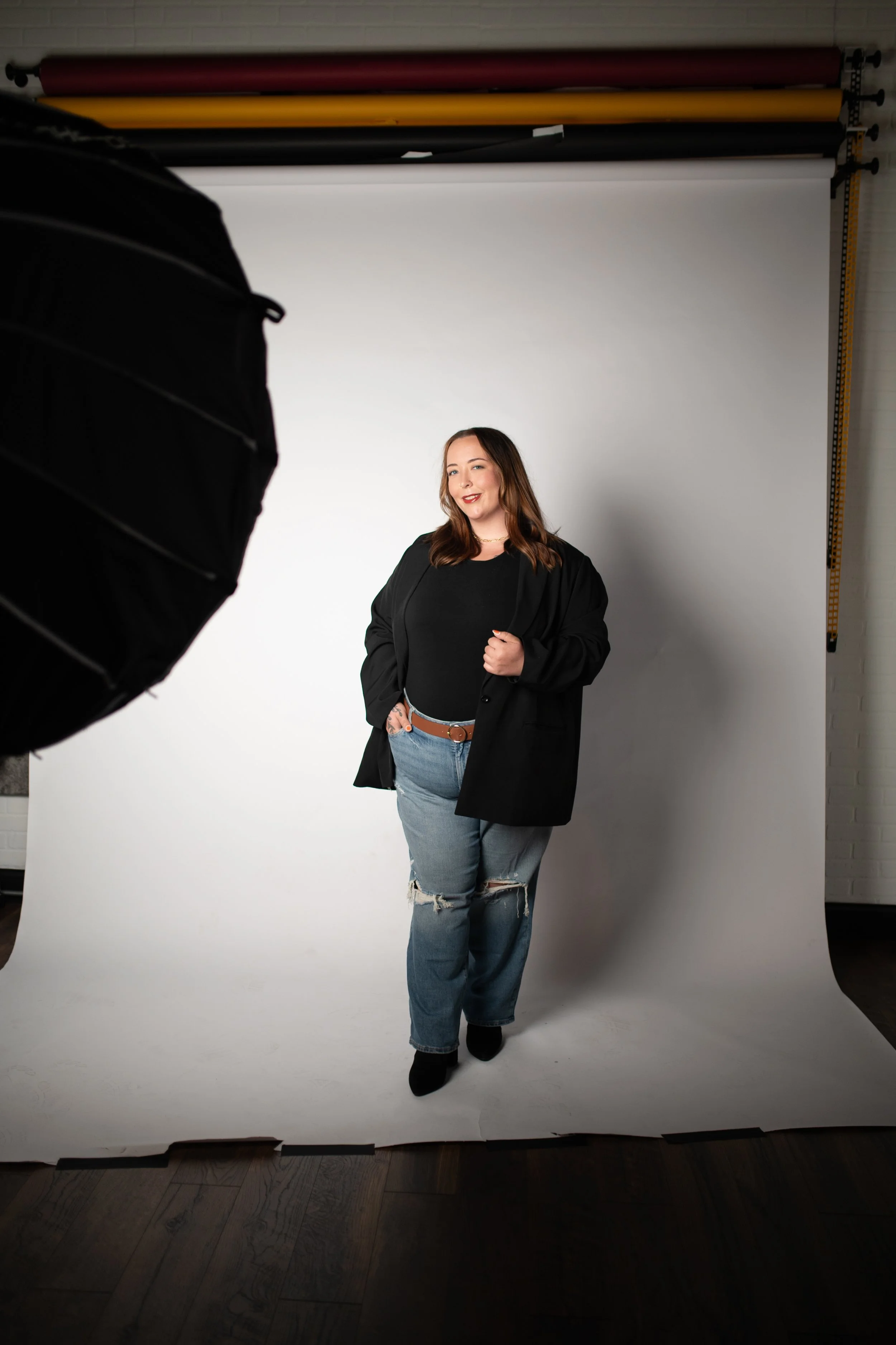 A woman with long brown hair, wearing a black jacket, black top, ripped jeans, and black shoes, posing in a photography studio with a white backdrop and professional lighting equipment near her.