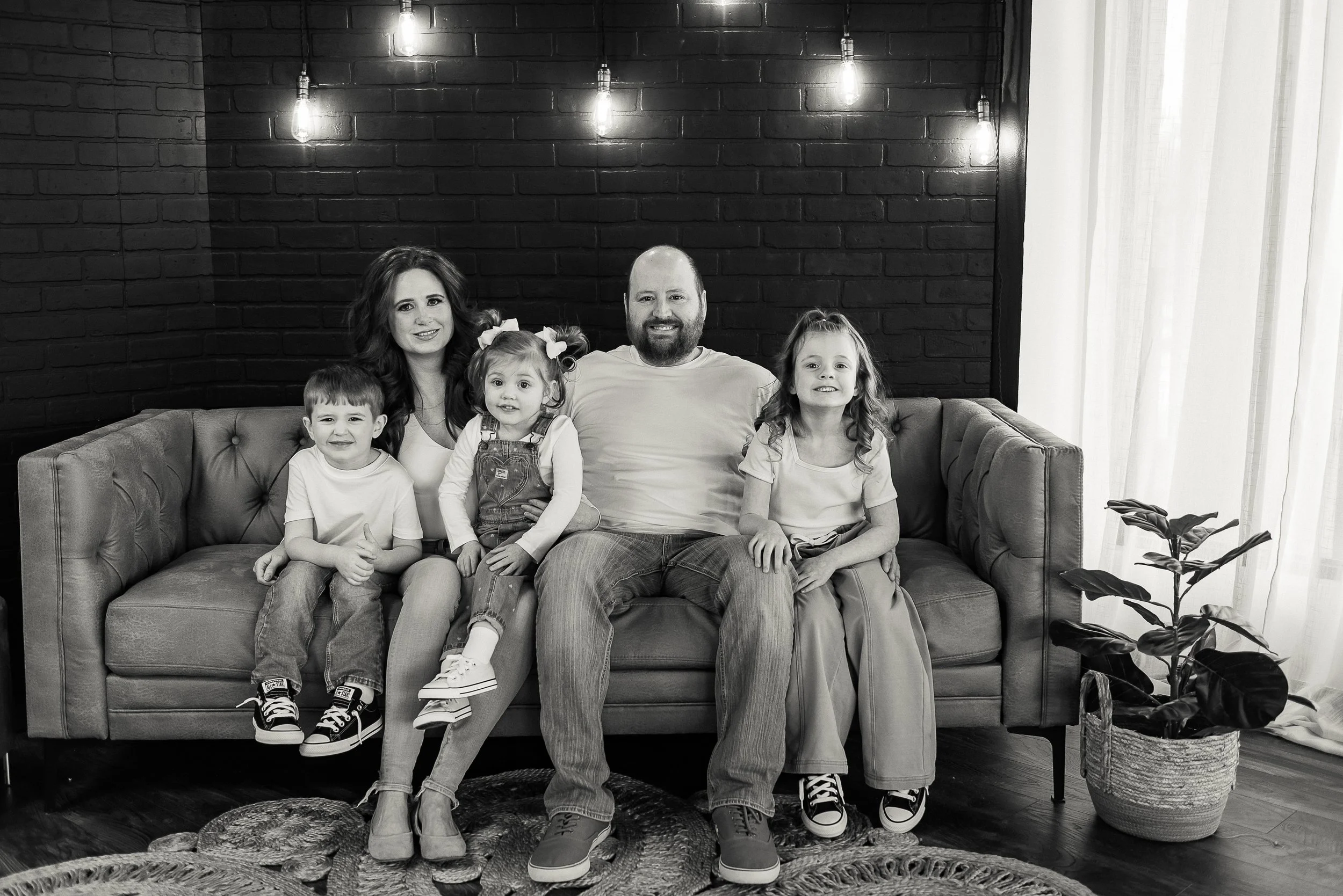 A black and white photo of a family of five sitting on a sofa in a modern room with a brick wall background and hanging light bulbs. The family includes two adults and three children, all smiling.