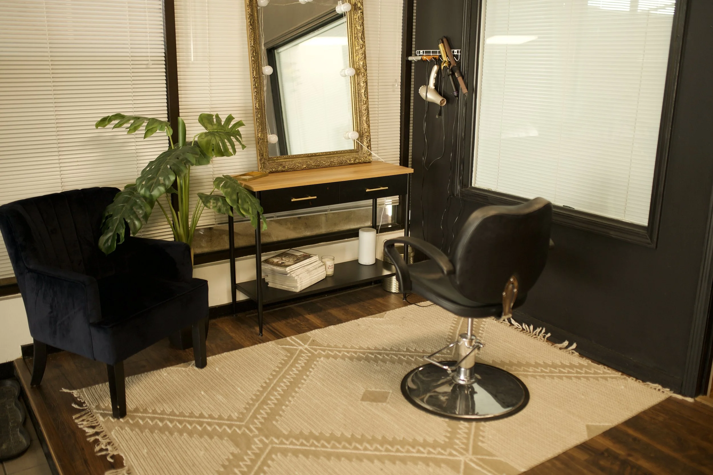 A salon or hair studio with a black salon chair, a black velvet armchair, a large green potted plant, a wooden table with a mirror, magazine stacks, a roll of paper towels, and a beige area rug on dark hardwood floors. The wall is painted dark, with windows covered by closed blinds.