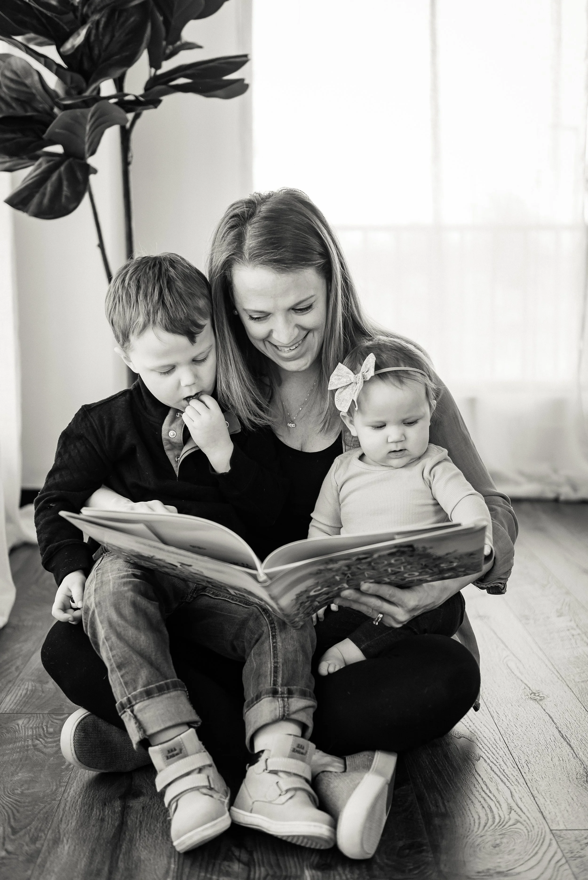 A woman and two children sitting on a wooden floor, reading a book together. The woman is smiling, the boy is looking at the book with curiosity, and the girl is also focused on the book, wearing a bow in her hair.