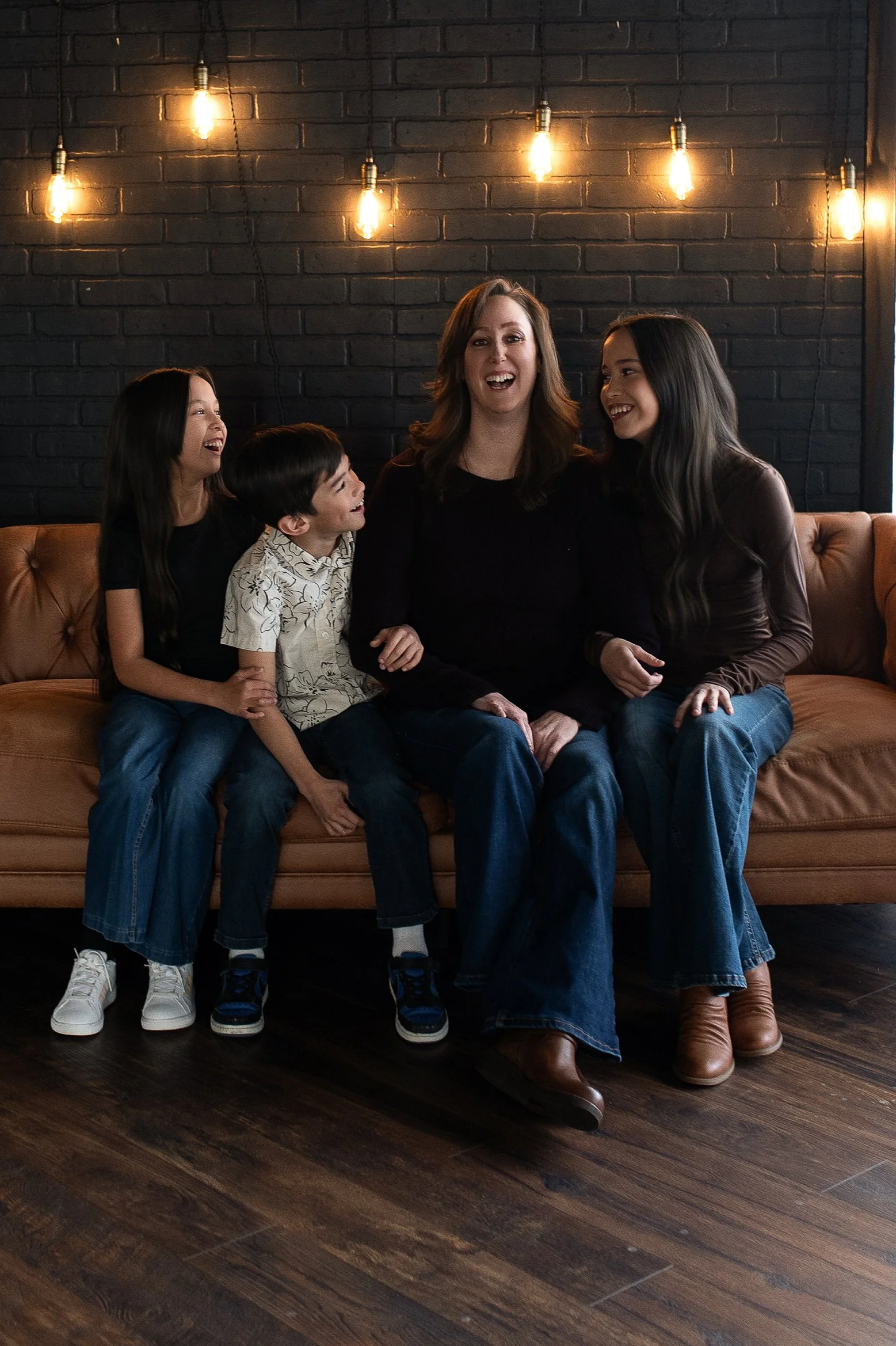 A group of four people sitting on a tan leather couch against a black brick wall with hanging Edison bulbs, smiling and enjoying each other's company.