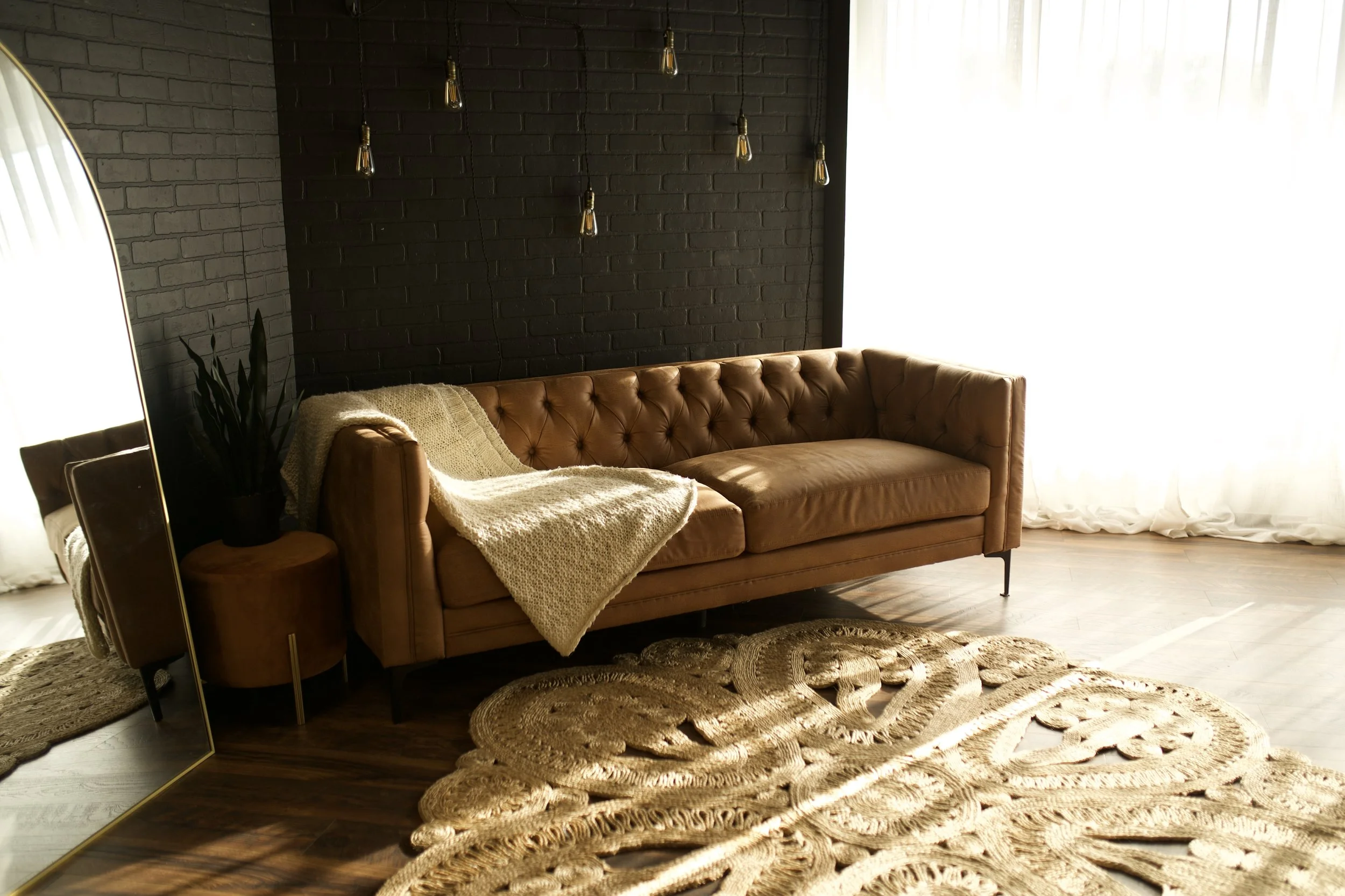 Living room with a tan leather tufted sofa, a cozy knit blanket, a large ornate beige rug, a full-length mirror, and natural light coming through sheer white curtains.