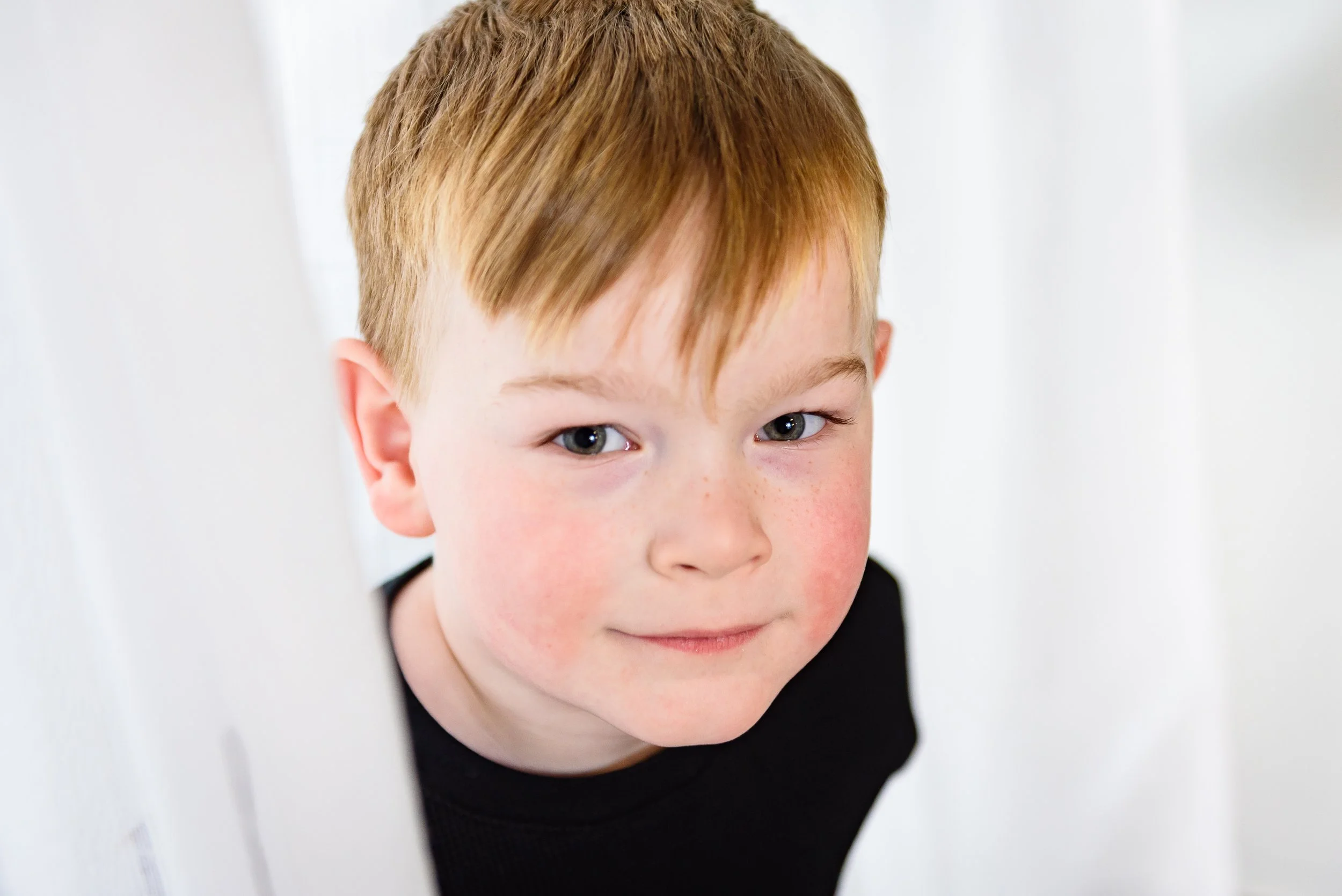 Close-up of a young boy with red hair and freckles peeking through a white doorframe, smiling softly.