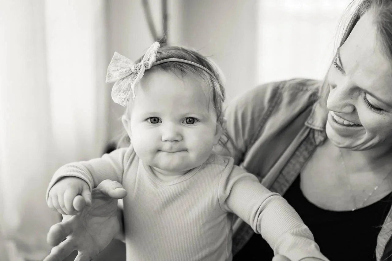 A woman with a smiling face holding a young girl who has a serious expression and wearing a bow headband.
