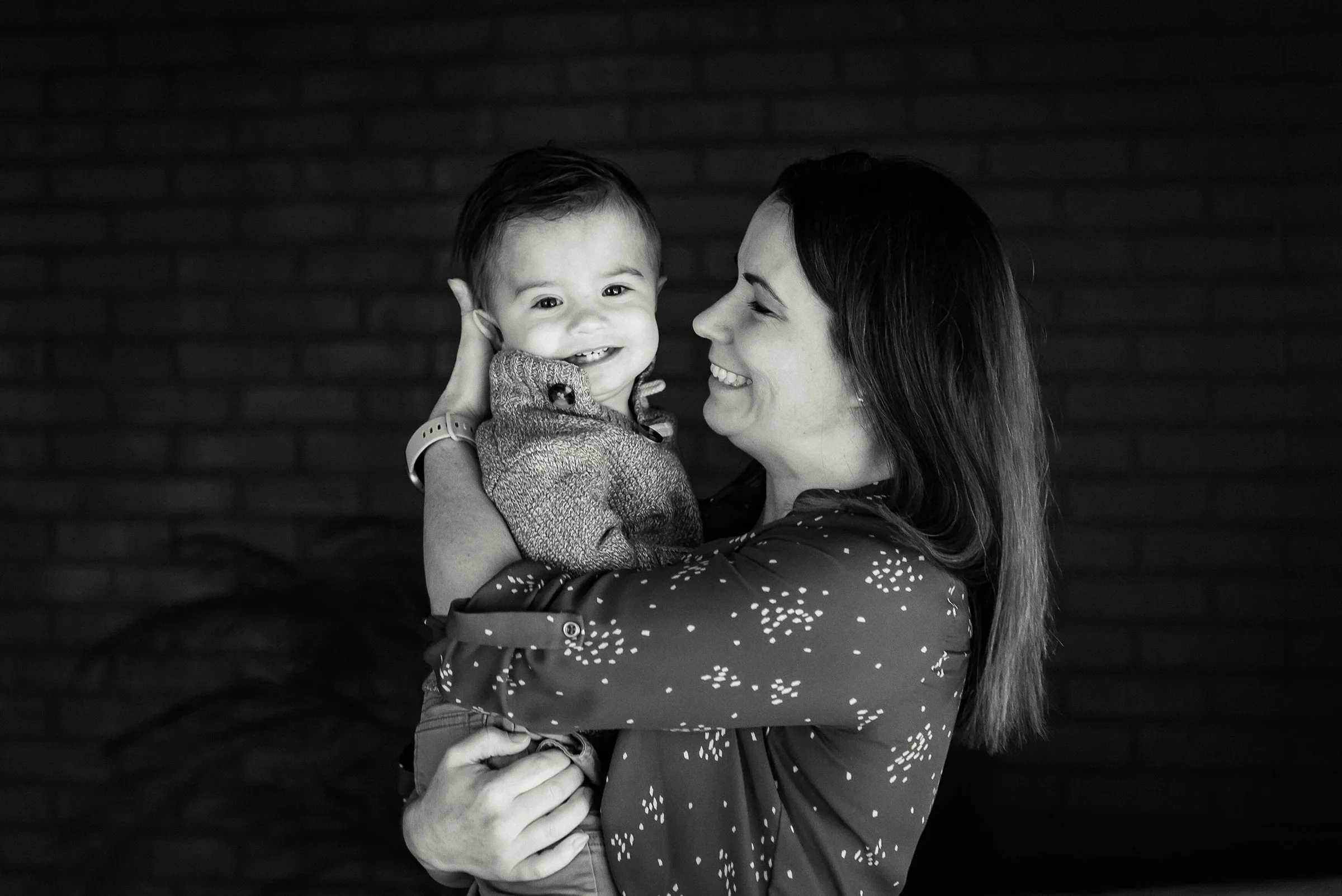 A woman holding a smiling young child in her arms, with a brick wall in the background, in black and white.