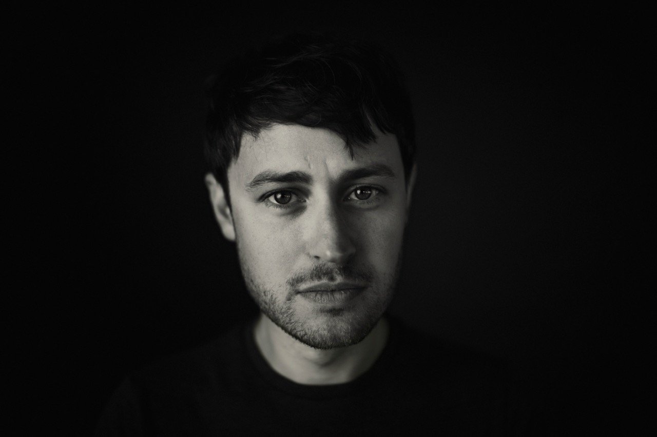 Black and white portrait of a young man with short dark hair, intense gaze, and slight facial hair, against a dark background.
