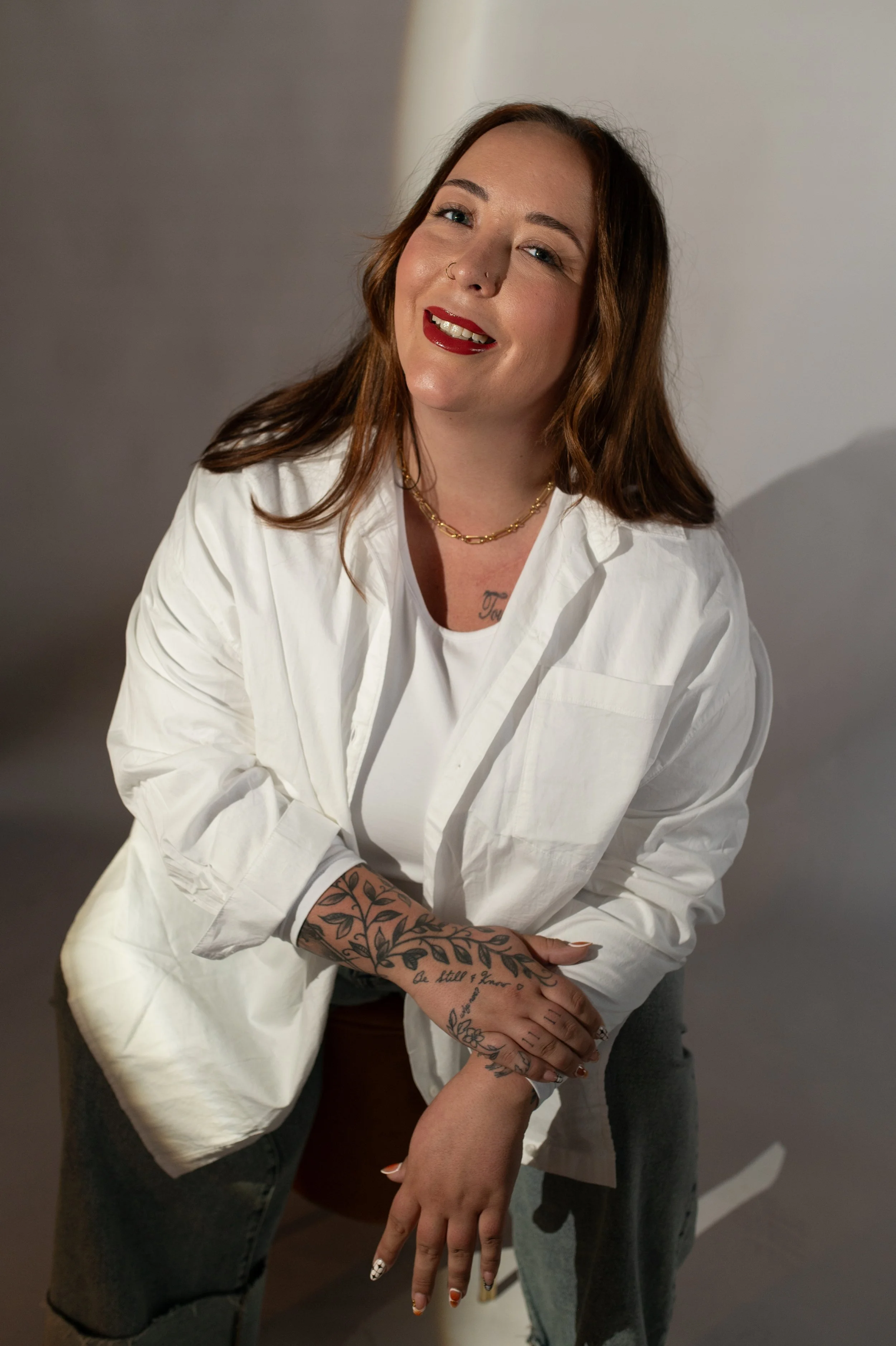A woman with long brown hair, red lipstick, and tattoos on her arms, smiling with her eyes closed. She is wearing a white shirt, gold necklace, and a nose ring, and is leaning against or interacting with another person in a casual setting.