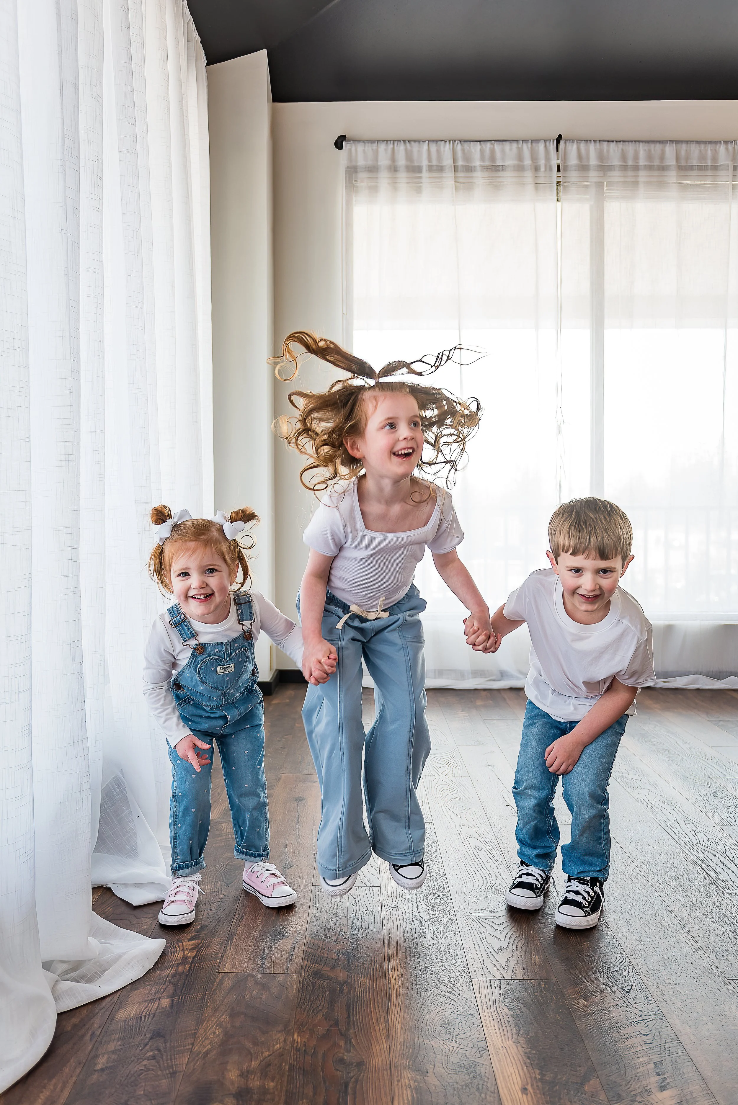 Three children holding hands and jumping in a bright room with white curtains and wooden floors.
