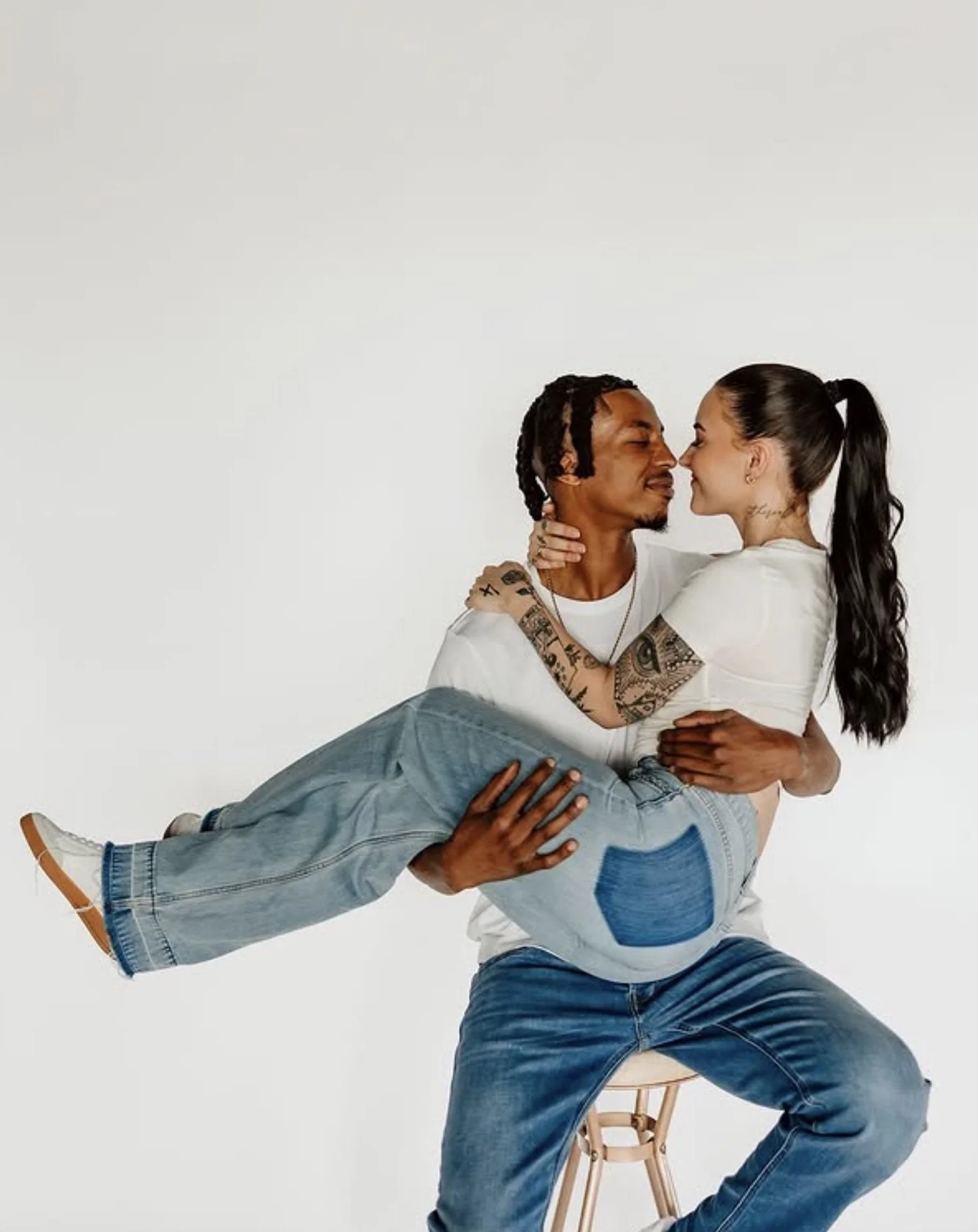 A man sitting on a wooden stool holding a woman in his lap. They are touching noses, looking at each other, and smiling. The man has tattoos and is wearing a white shirt and blue jeans. The woman has long dark hair in a high ponytail, tattoos on her 