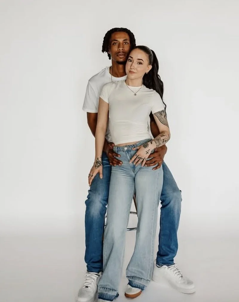 A young couple standing close together against a plain white background, with the man behind the woman, both looking directly at the camera.