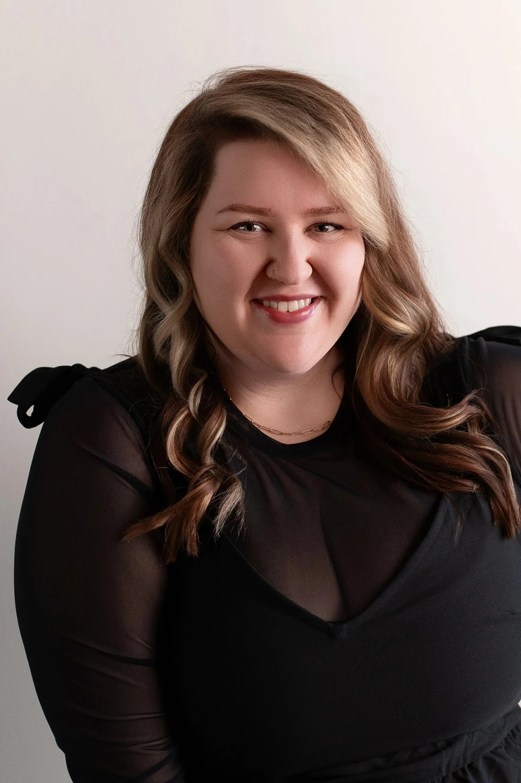 A woman with light brown, wavy hair, wearing a black sheer top, smiling, in front of a plain background.