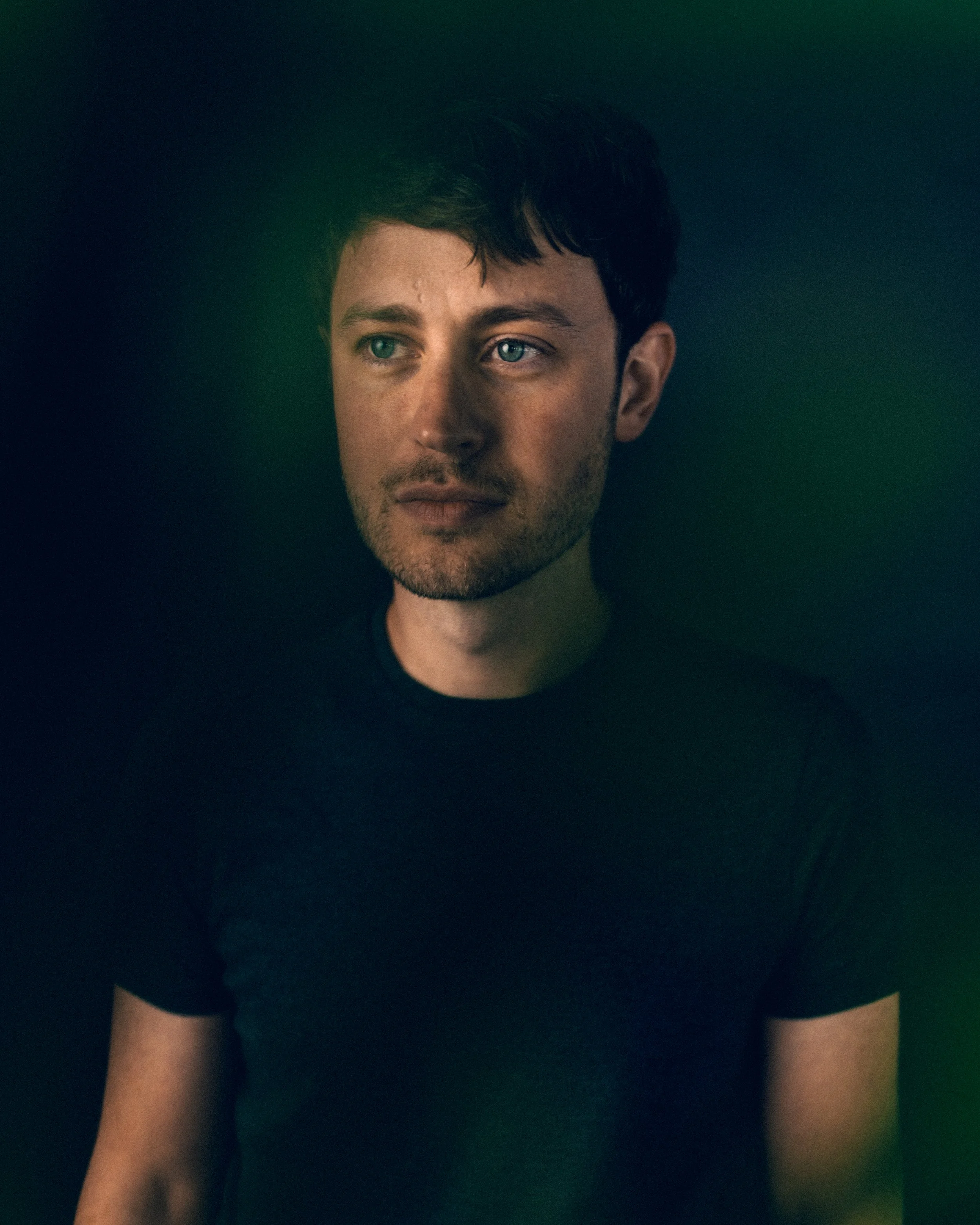 A young man with blue eyes and dark hair, wearing a black shirt, is looking off to the side. The background has a dark gradient with a hint of green.