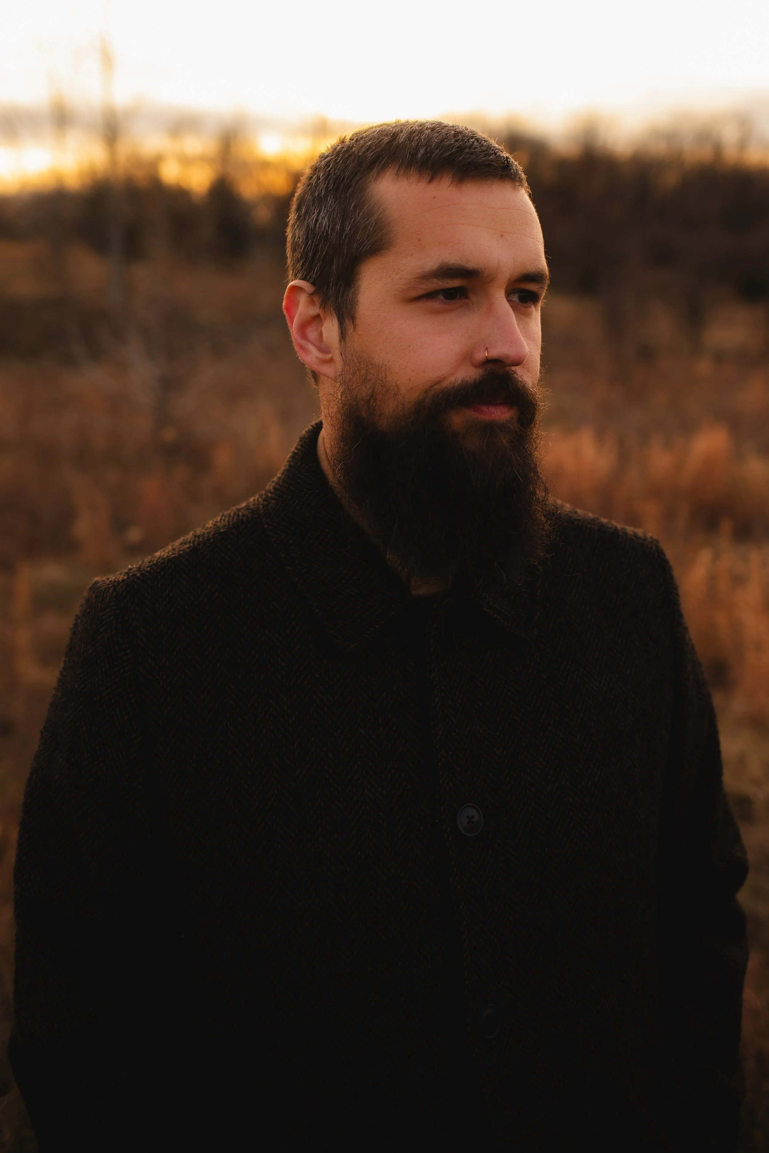 A man with a beard and mustache, wearing a dark jacket, standing outdoors during sunset with a blurry background of trees and plants.