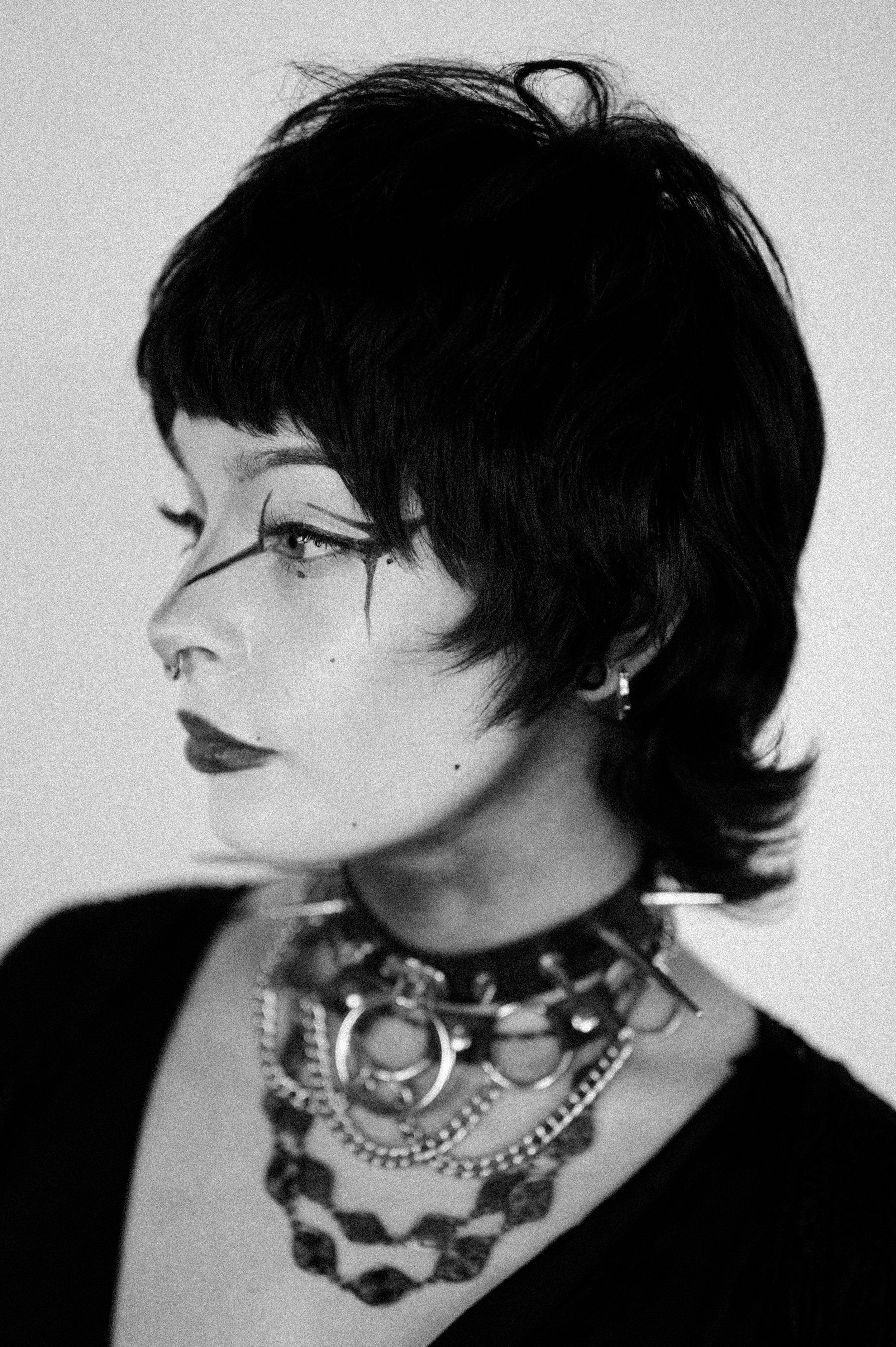 Black and white portrait of a woman with short, layered hair and bold makeup, wearing multiple necklaces and earrings, with a dark top.