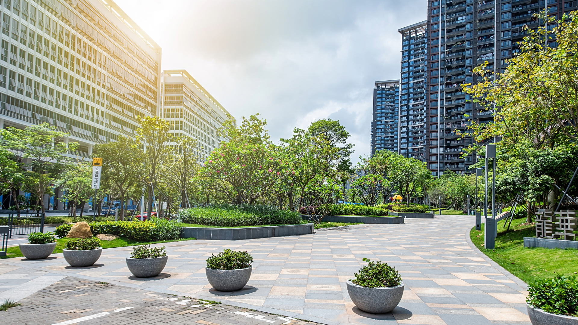 A city park with paved walkways, green trees, shrubs, and potted plants, surrounded by tall modern buildings with glass windows, under a partly cloudy sky with sunlight.
