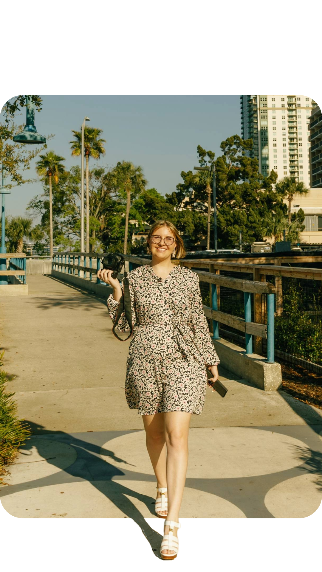 A young woman wearing glasses and a floral dress, holding a camera and a phone, walks on a sunny sidewalk with trees and buildings in the background.