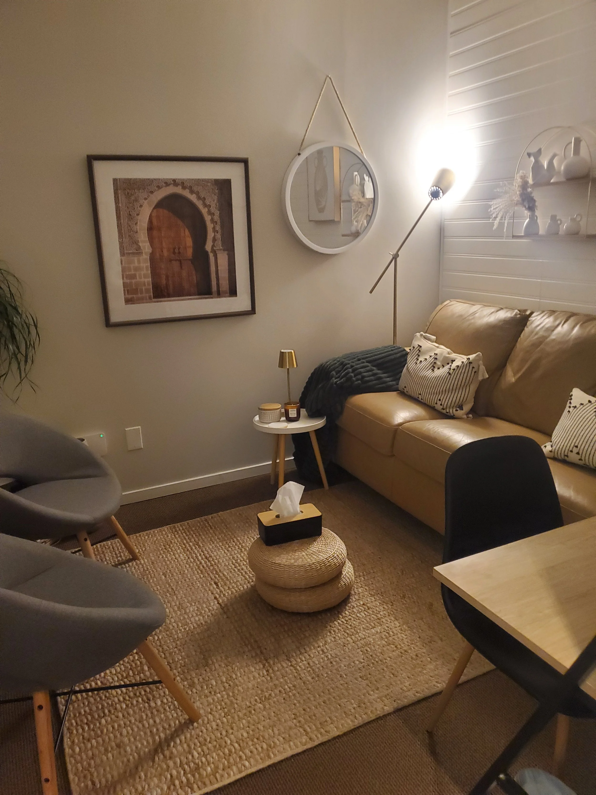 A cozy living room with a beige leather sofa, a black armchair, a wooden dining table, and a small round side table with a gold lamp. The wall features framed artwork, a round mirror, and a white shelf with decorative vases. There is a floor lamp and a woven rug on the floor.
