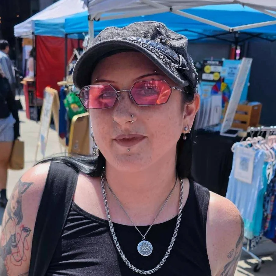 A woman with tattoos, piercings, pink mirrored sunglasses, and a gray cap, standing in an outdoor marketplace with tents and clothing racks in the background.