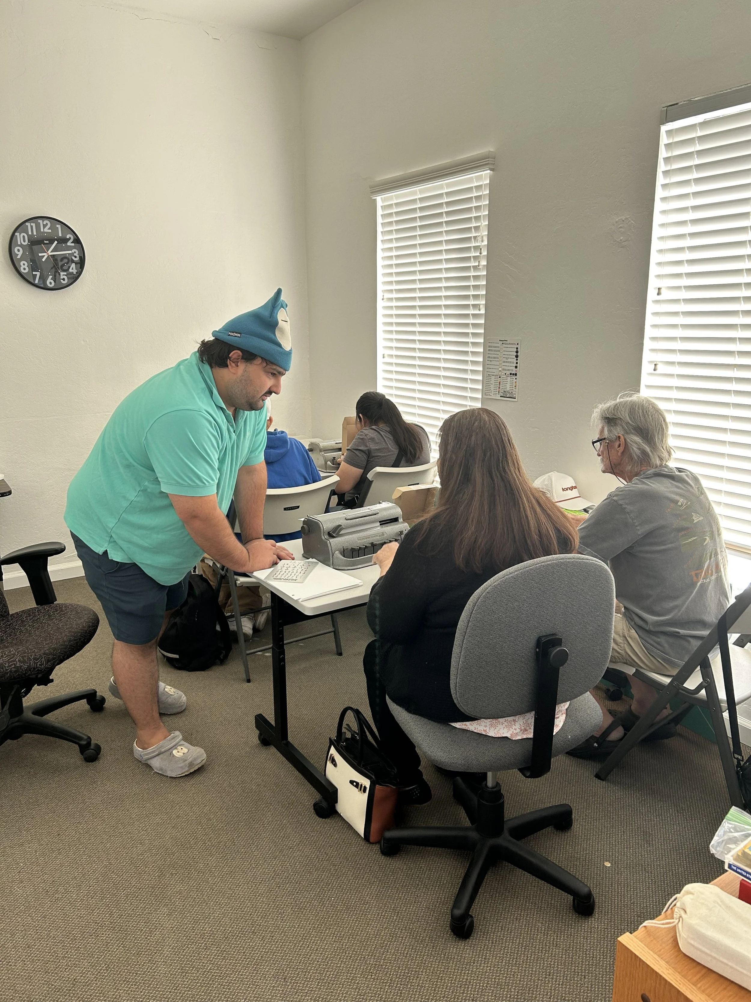 Braille instructor Jacob Gaddam works with students during a Beginning Braille class at VIPS, demonstrating Braille on a paper worksheet beside a Braille typewriter while students practice at their desks.