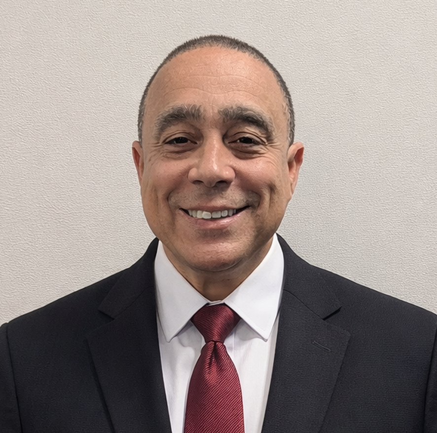 A man in a black suit with a white shirt and a red tie, smiling in front of a plain wall.