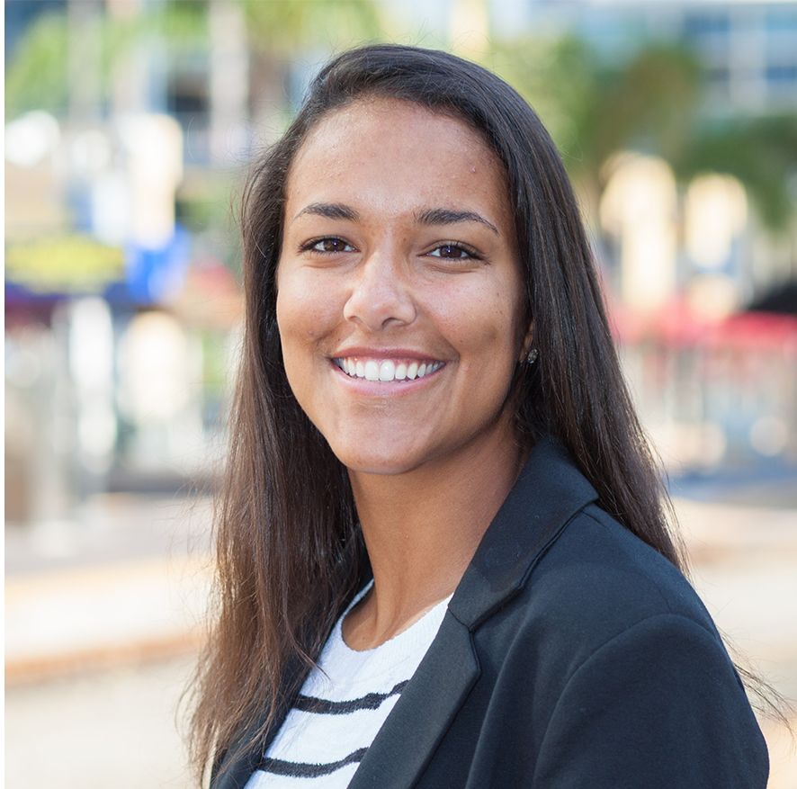 A young woman with long dark hair smiling outdoors, wearing a navy blazer and striped shirt, with blurred trees and buildings in the background.