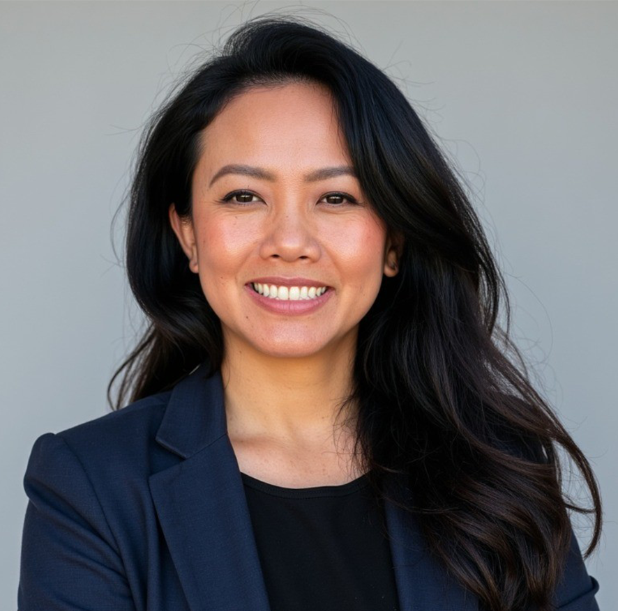 A woman with long black hair smiling, wearing a navy blazer and black top, standing against a plain background.