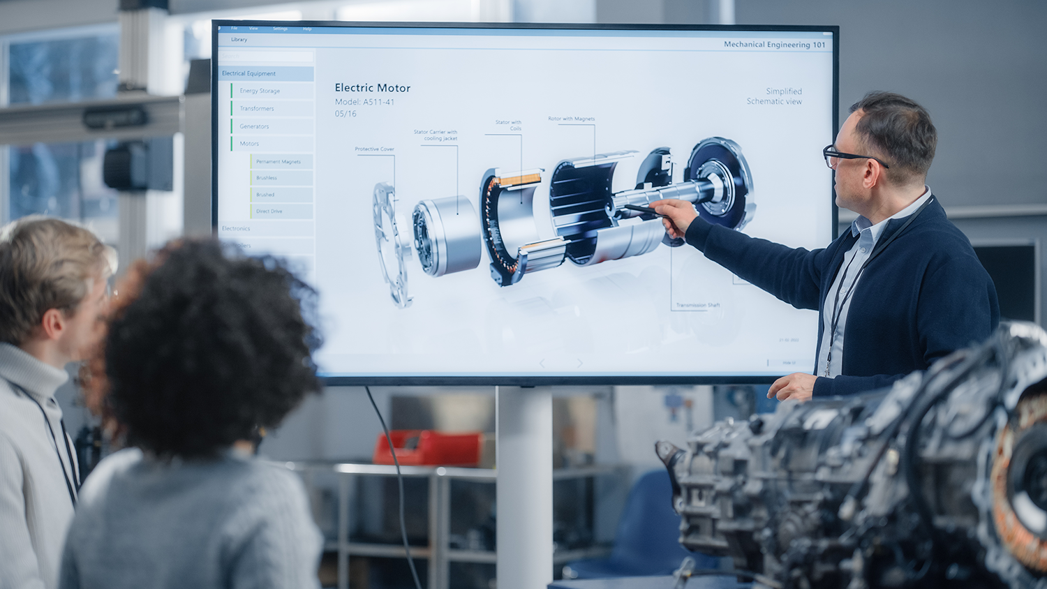 A man in glasses and a dark blazer is giving a presentation about an electric motor diagram on a large screen in a classroom or laboratory setting, with students observing.