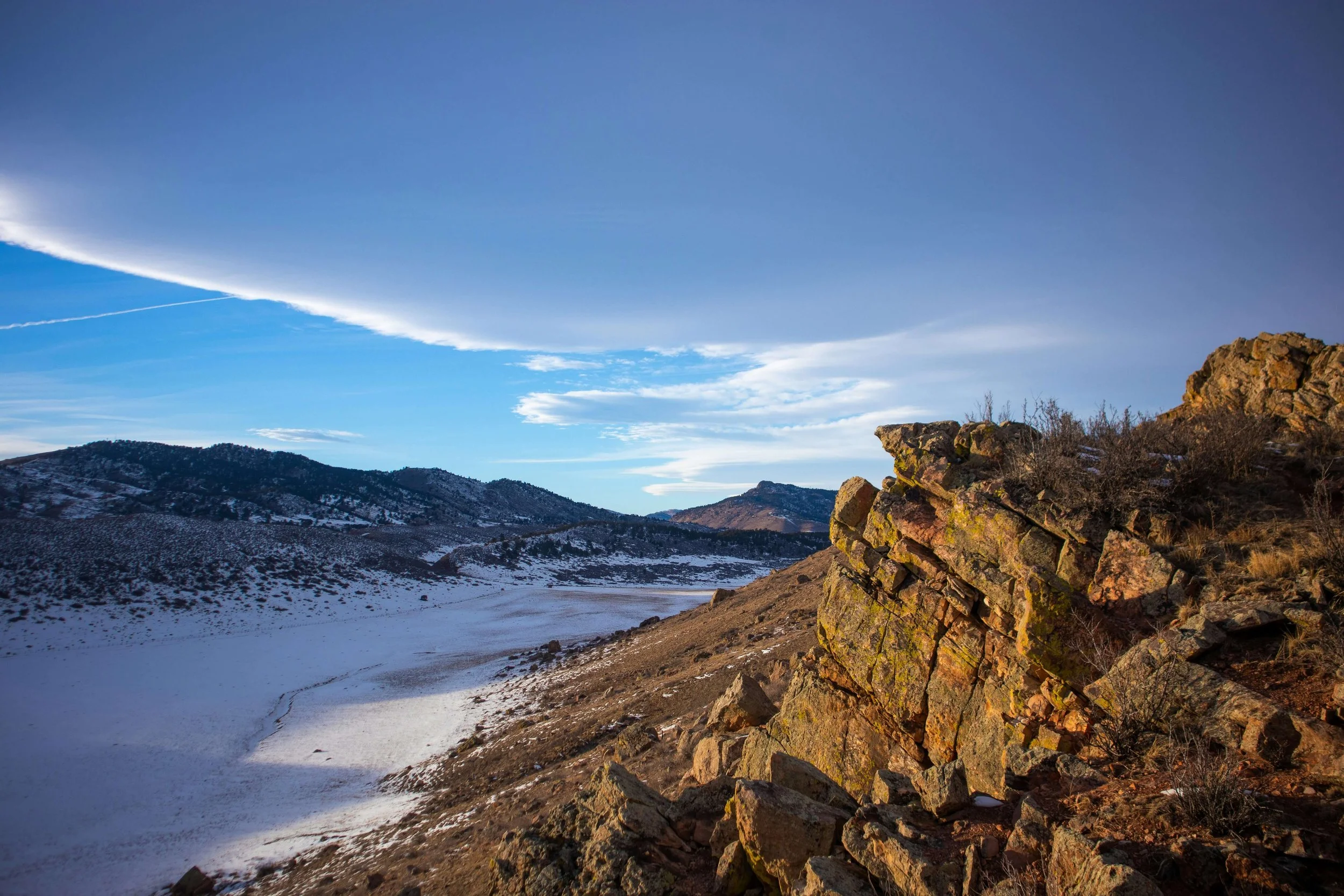 Scenic mountain landscape with snow-covered ground, rocky hillside, and hills in the distance under a mostly clear sky with some clouds.