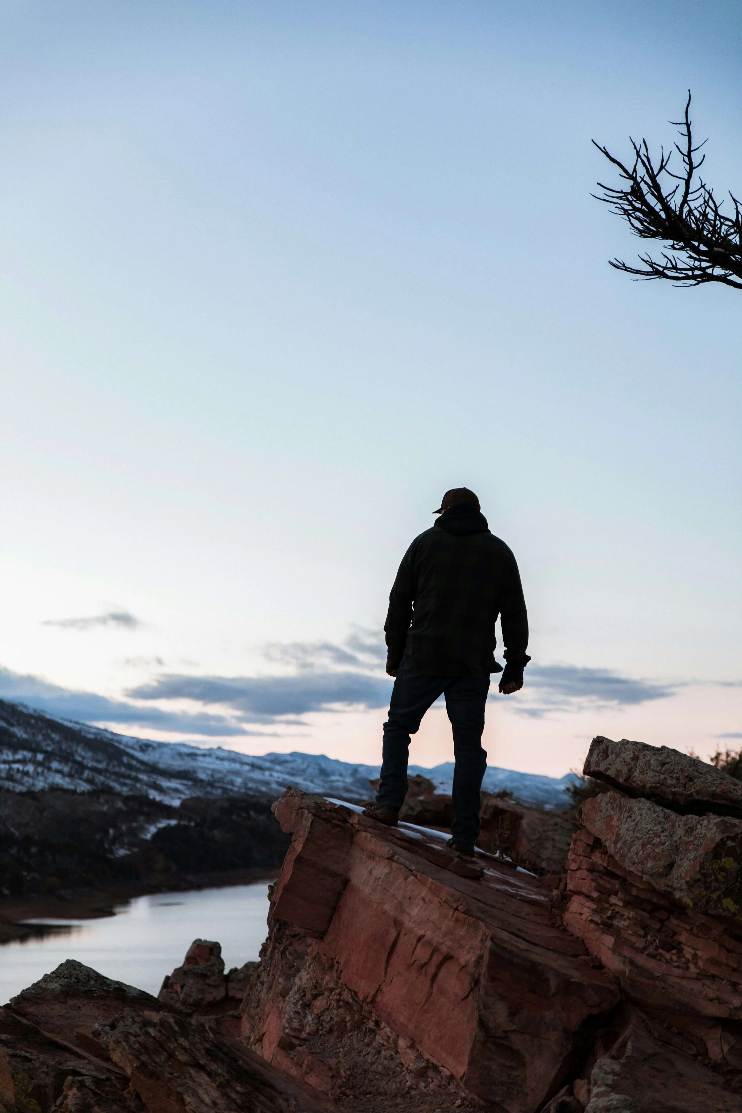 A man standing on a large fallen tree trunk in a mountainous, snowy landscape during twilight, with mountains and a cloudy sky in the background.