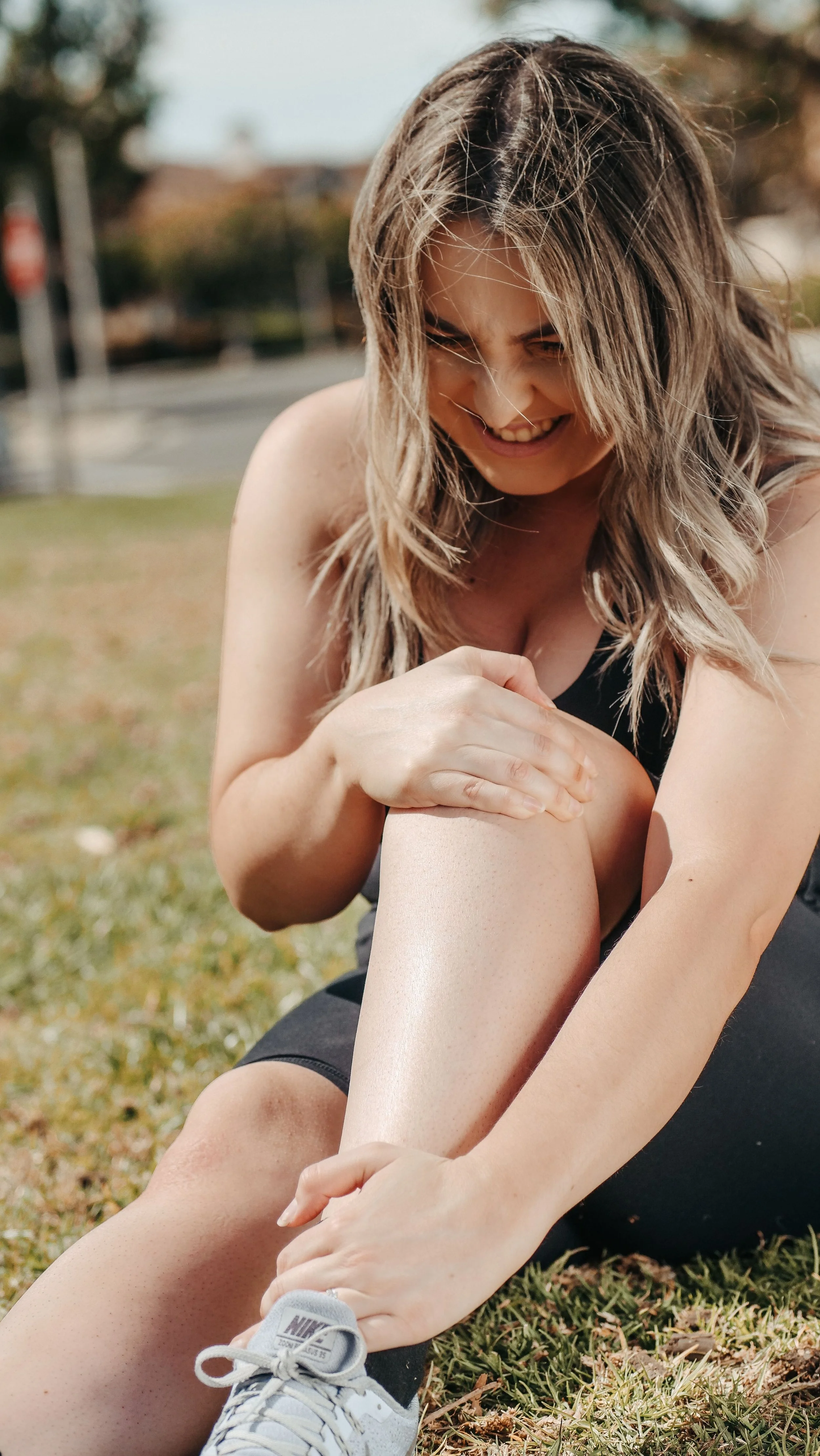 A young woman with long, wavy hair smiling and stretching outside on grass, wearing black athletic clothing and gray athletic shoes.