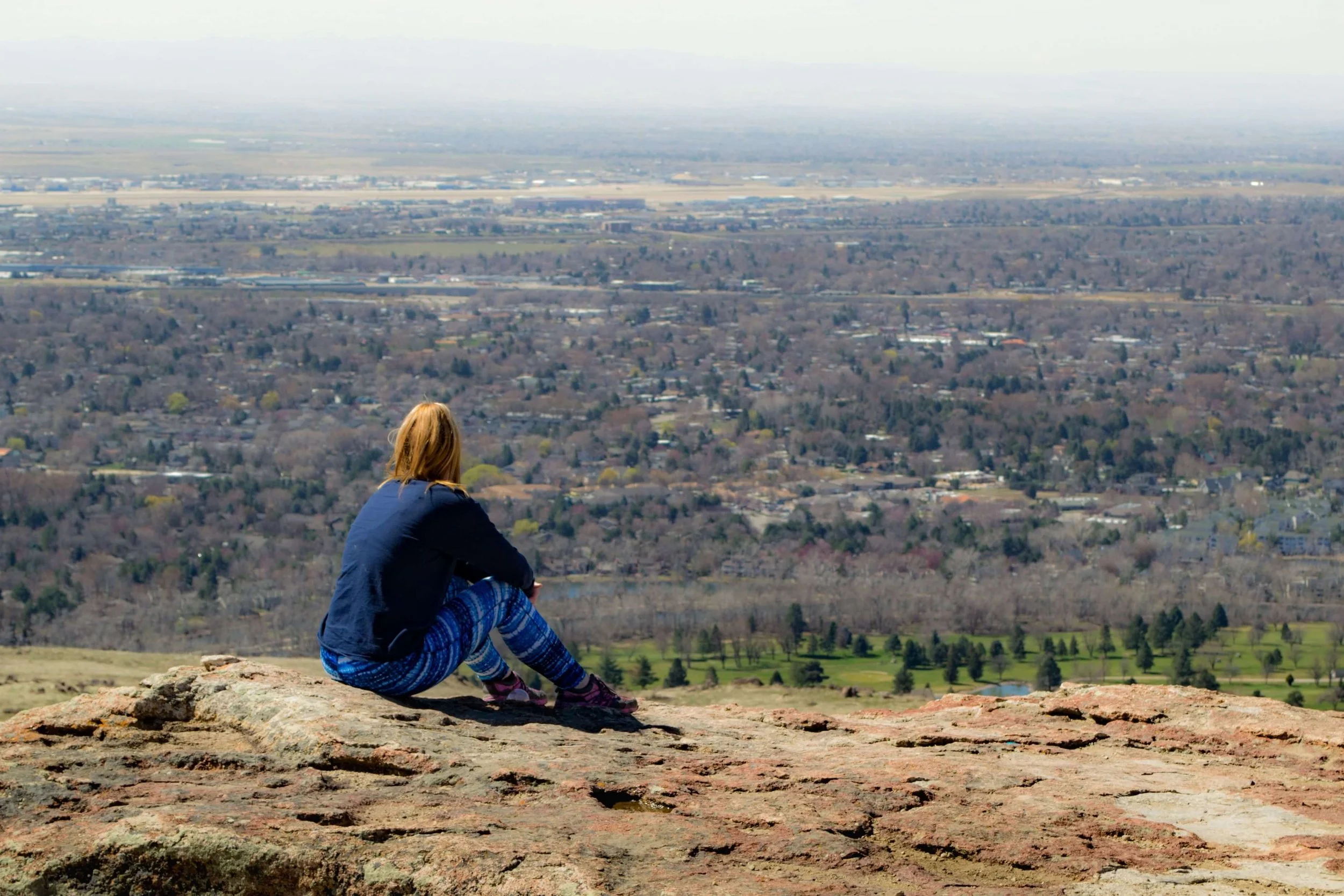Person with blond hair sitting on rocky ground, overlooking a cityscape and landscape, wearing a blue jacket and patterned leggings.