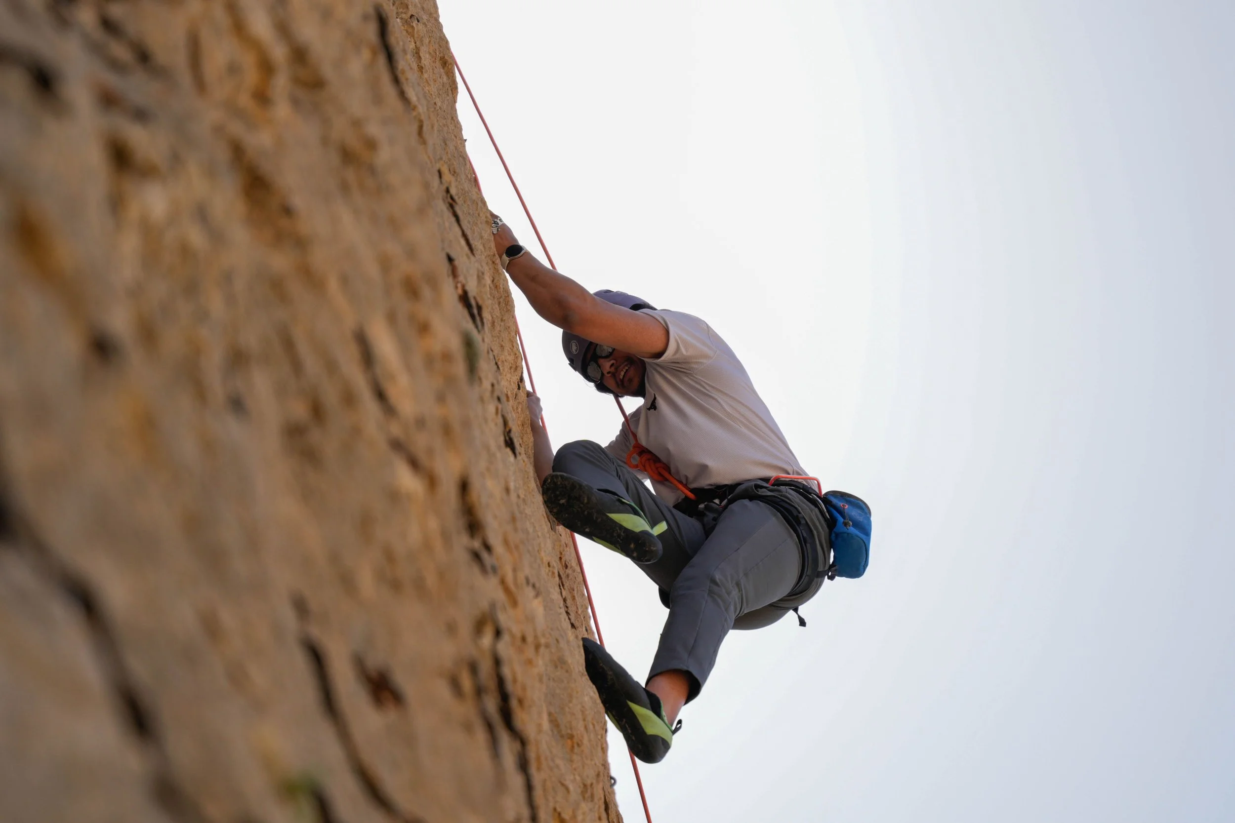 A person wearing a helmet and climbing gear is ascending a rocky cliff face, holding a rope and smiling, with a clear sky in the background.