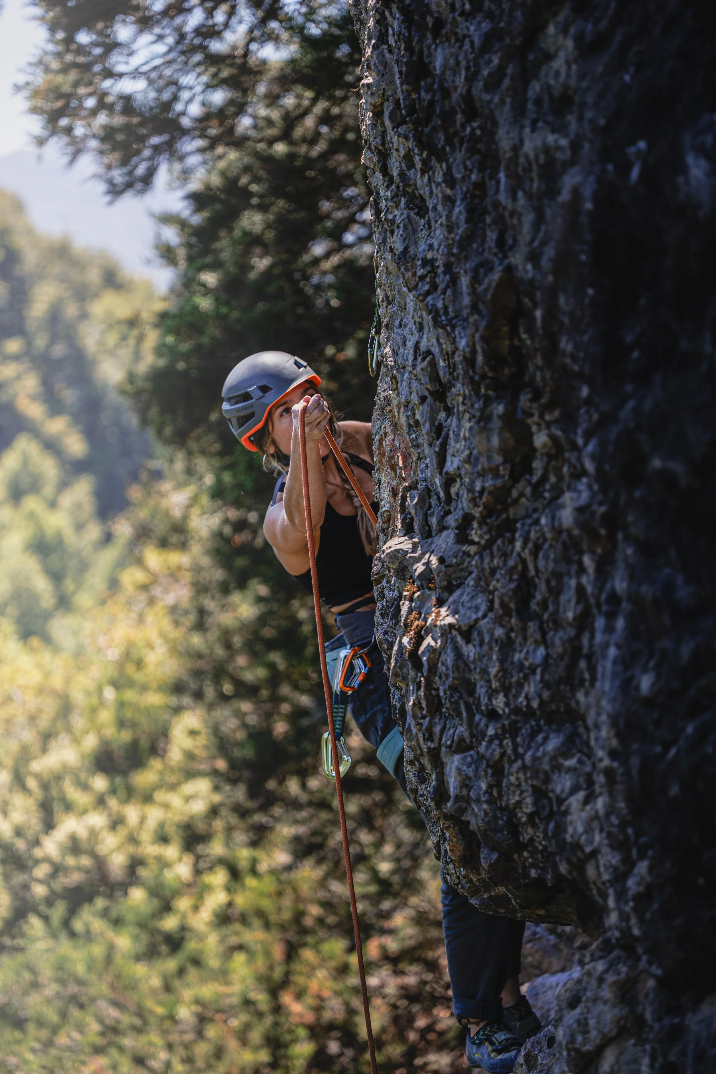 A woman rock climbing outdoors, wearing a helmet, using a harness, and holding a climbing rope.
