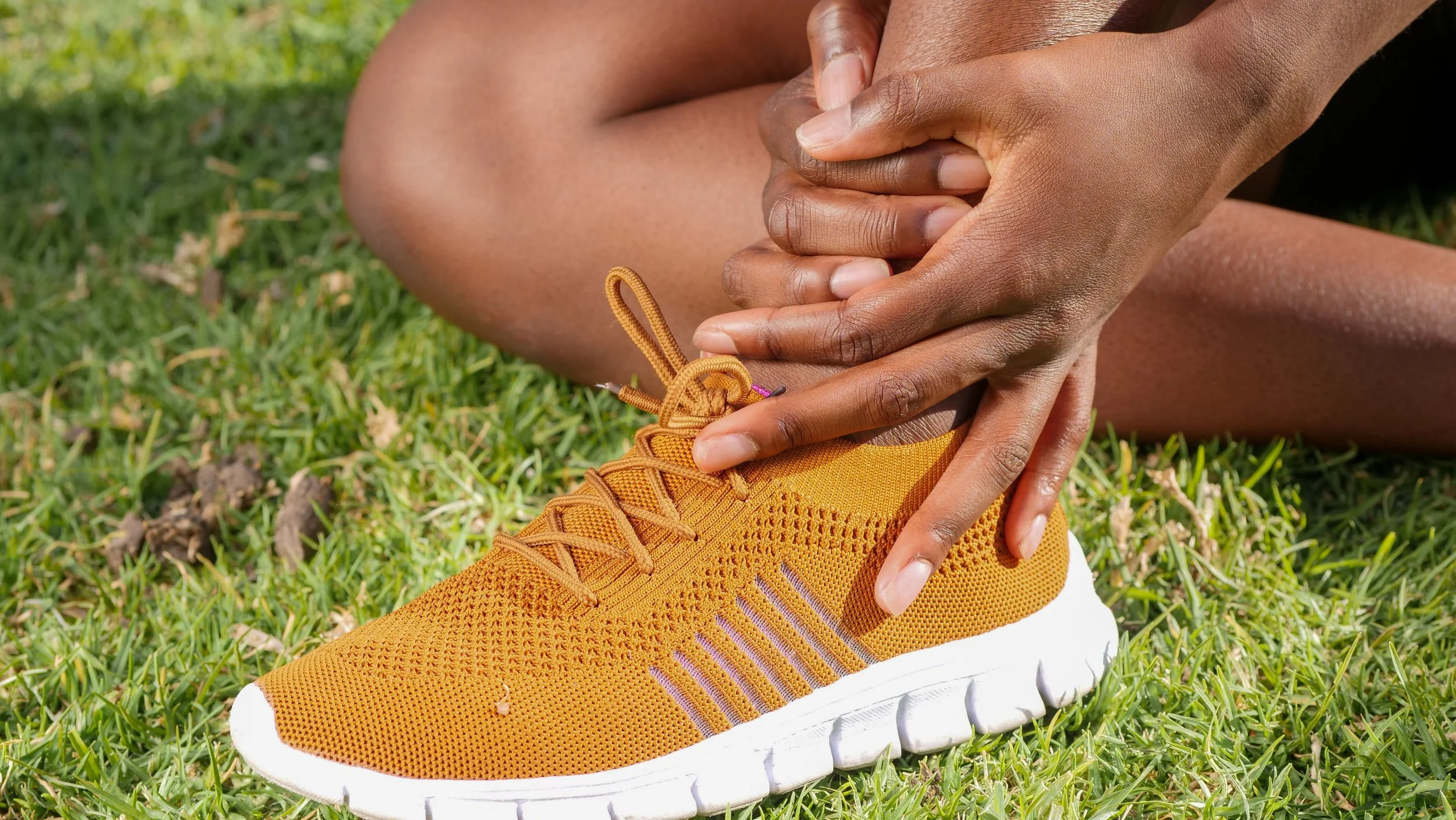 Person tying their teal shoelace on an orange athletic shoe while sitting on green grass.
