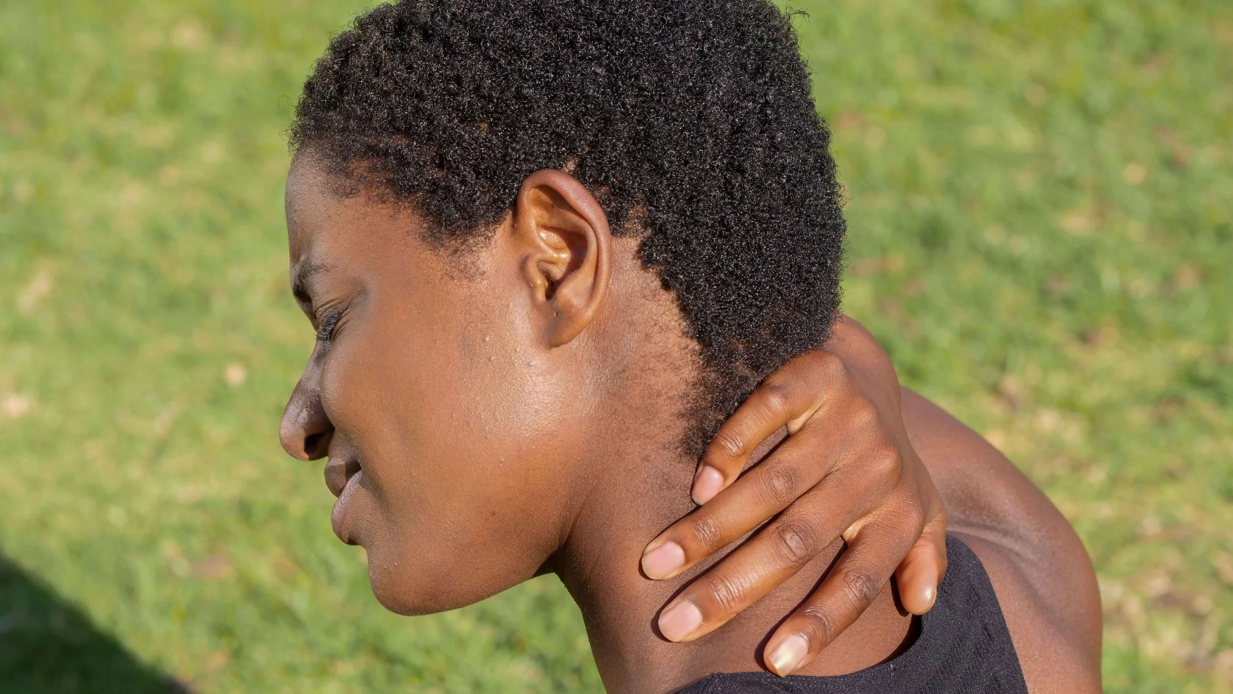 A person with short, curly black hair holding their neck outdoors with a green grass background.