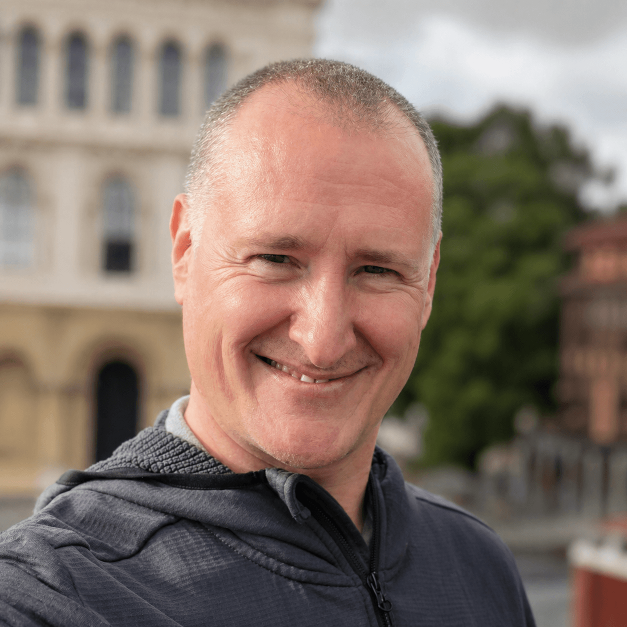 Smiling man with short hair wearing a dark jacket outdoors, with historic buildings and green trees in the background.